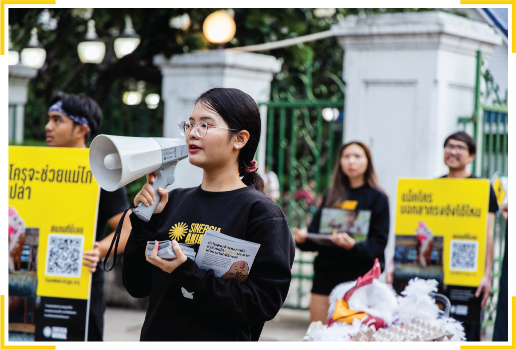 A close-up of a young female Sinergia Animal activist speaking into a megaphone and holding campaign leaflets during an outdoor street action in Thailand