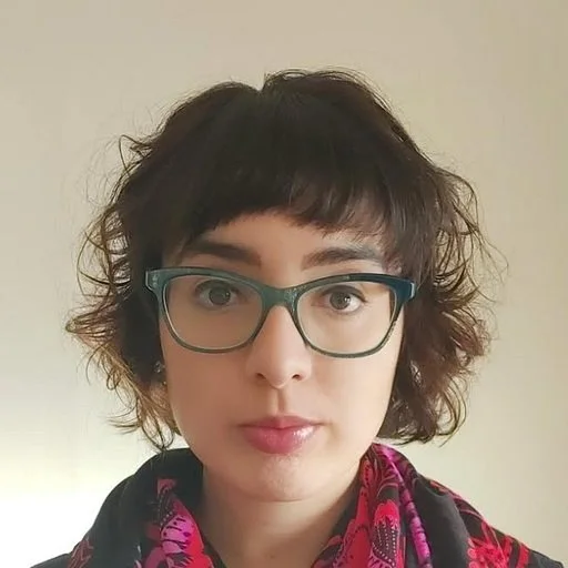 A woman with short, curly dark hair and glasses looks into the camera. She is wearing a black top and a colorful pink and black patterned scarf. The background is a plain, off-white wall.