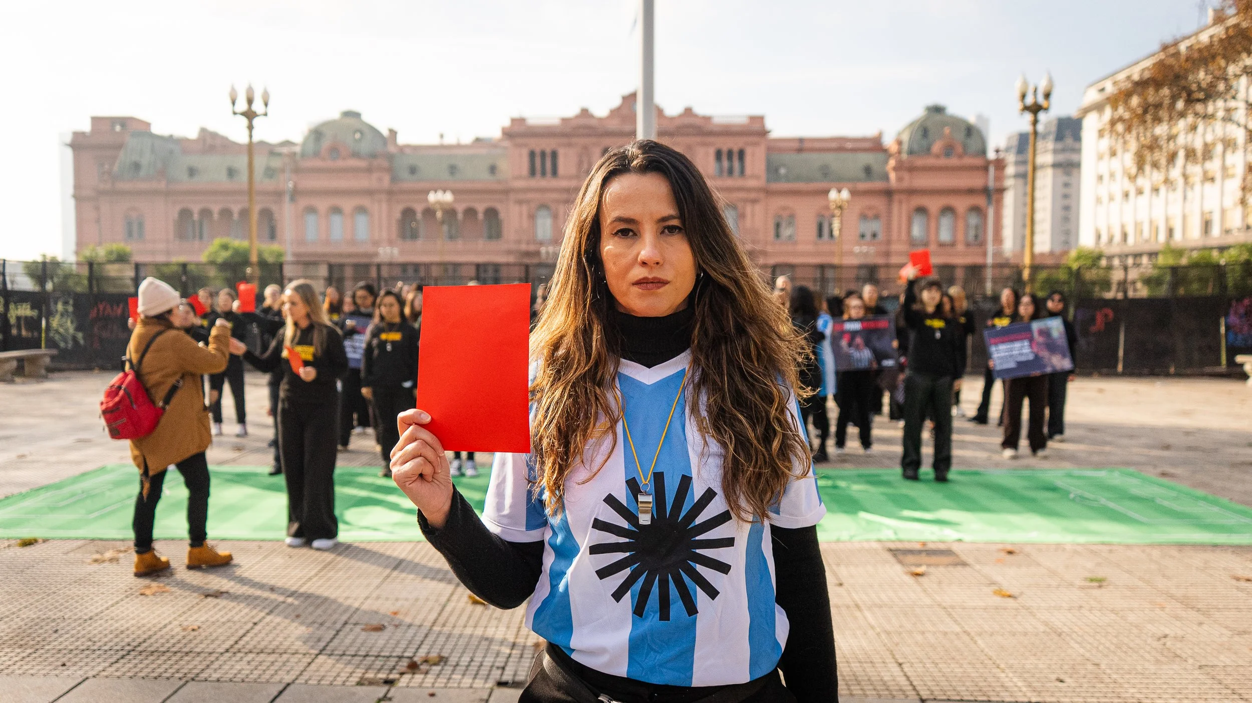Carolina Galvani, Executive Director of Sinergia Animal, stands in Plaza de Mayo wearing an Argentina-style blue and white striped shirt and holding up a red card. Other activists and the Casa Rosada building are visible in the background.
