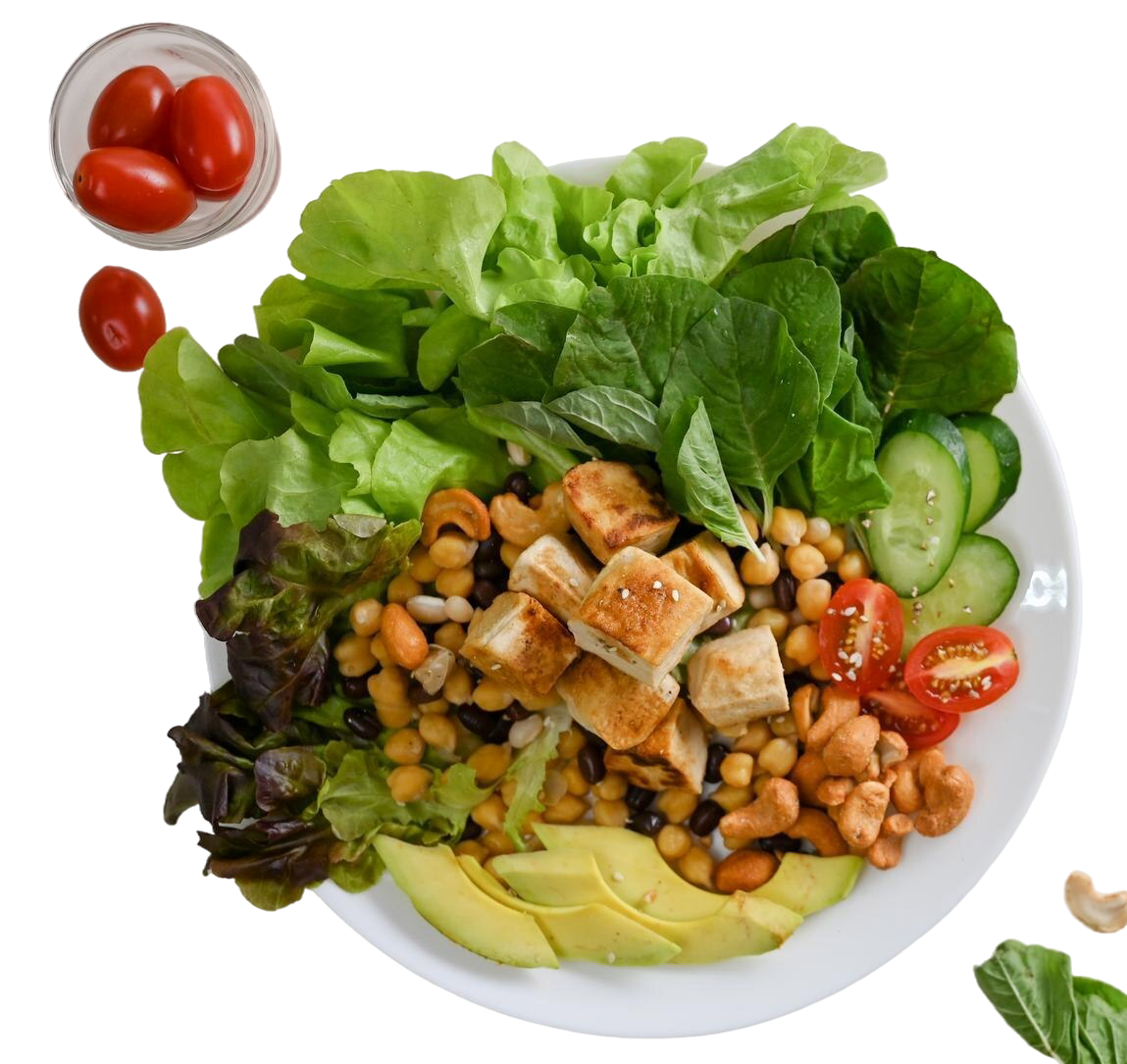 A white bowl filled with a mixed salad containing lettuce, spinach, cucumber slices, cherry tomatoes, avocado slices, chickpeas, black beans, grilled chicken pieces, and crispy chickpeas, with a small bowl of cherry tomatoes nearby.