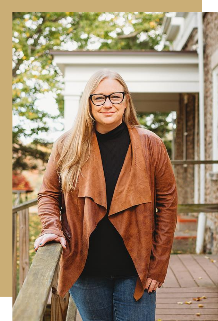 A woman with long blonde hair, glasses, and a black top, wearing a brown leather jacket, leaning on a wooden railing outside, with a house and trees in the background.