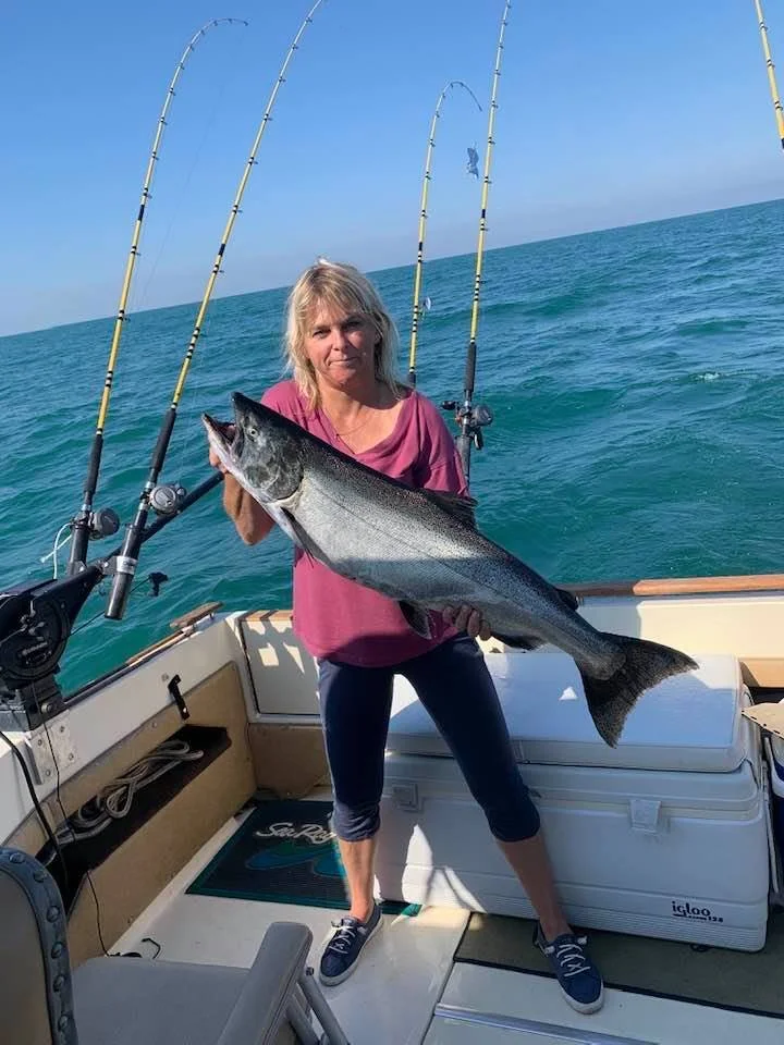 A woman on a boat holding a large fish she caught during a fishing trip, with blue ocean water and three fishing rods in the background.