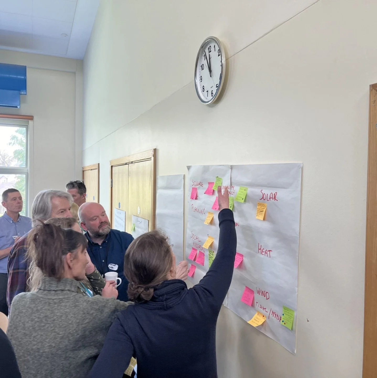 A group of people at Food and Farming Climathon. They are gathered in a room participating in a discussion around a large sheet of paper on the wall with colorful sticky notes. A woman is pointing at the sticky notes while others observe.