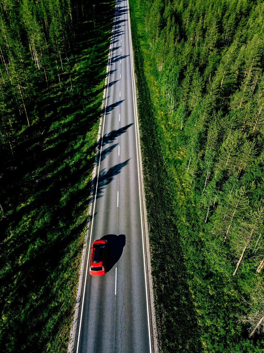 An aerial view of a red car driving on a two-lane road through a dense green forest.