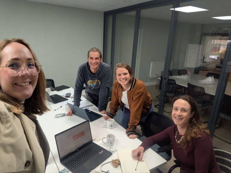 Members of FODCAP staff in a meeting. Four people in a meeting room smiling around a table with laptops, mugs, and notebooks, with a glass wall showing another room in the background.