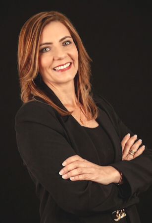 Professional woman with brown hair, smiling, wearing a black blazer, crossing her arms against a black background.