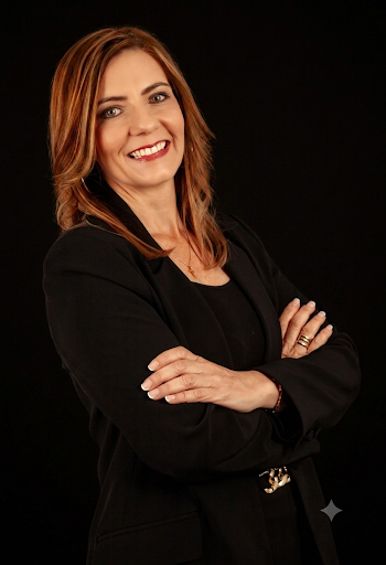 A woman with shoulder-length brown hair wearing a black blazer and smiling against a black background.