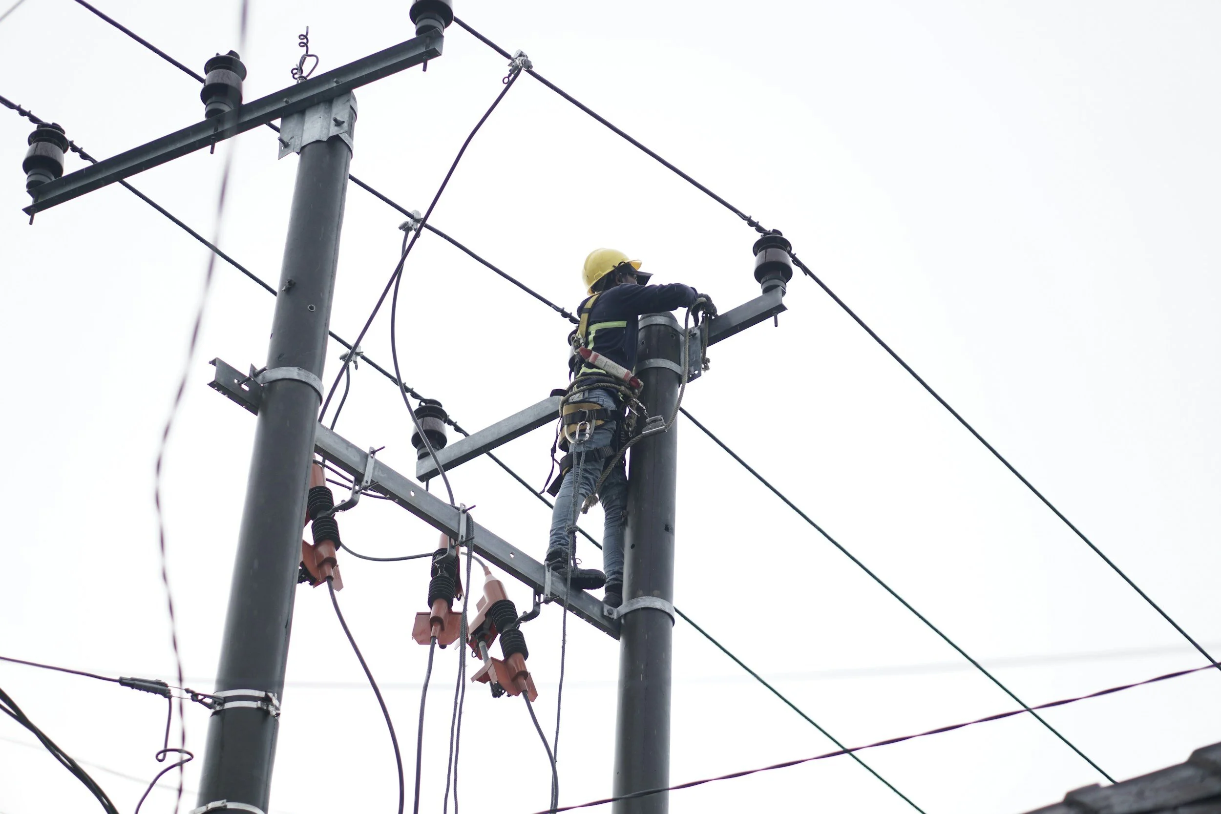 A utility worker in a safety harness performs maintenance on a power line atop a utility pole.