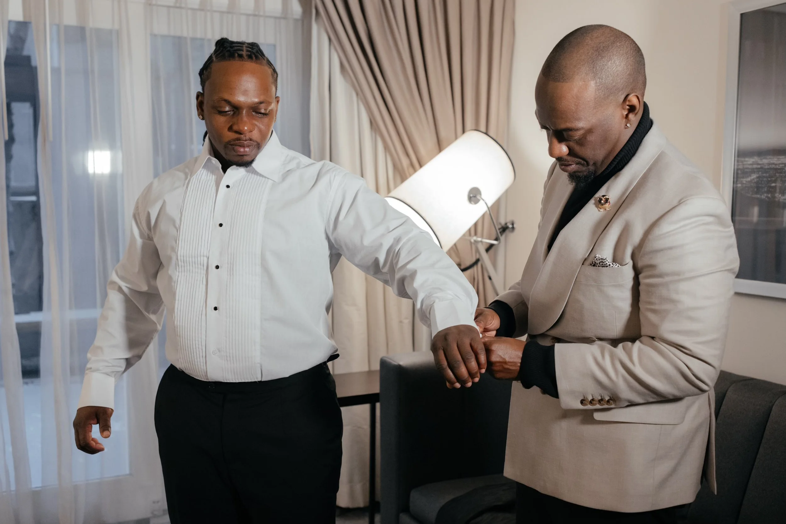 Two men in formal attire, one adjusting his watch while the other looks on, standing in a well-lit room with curtains and a lamp.