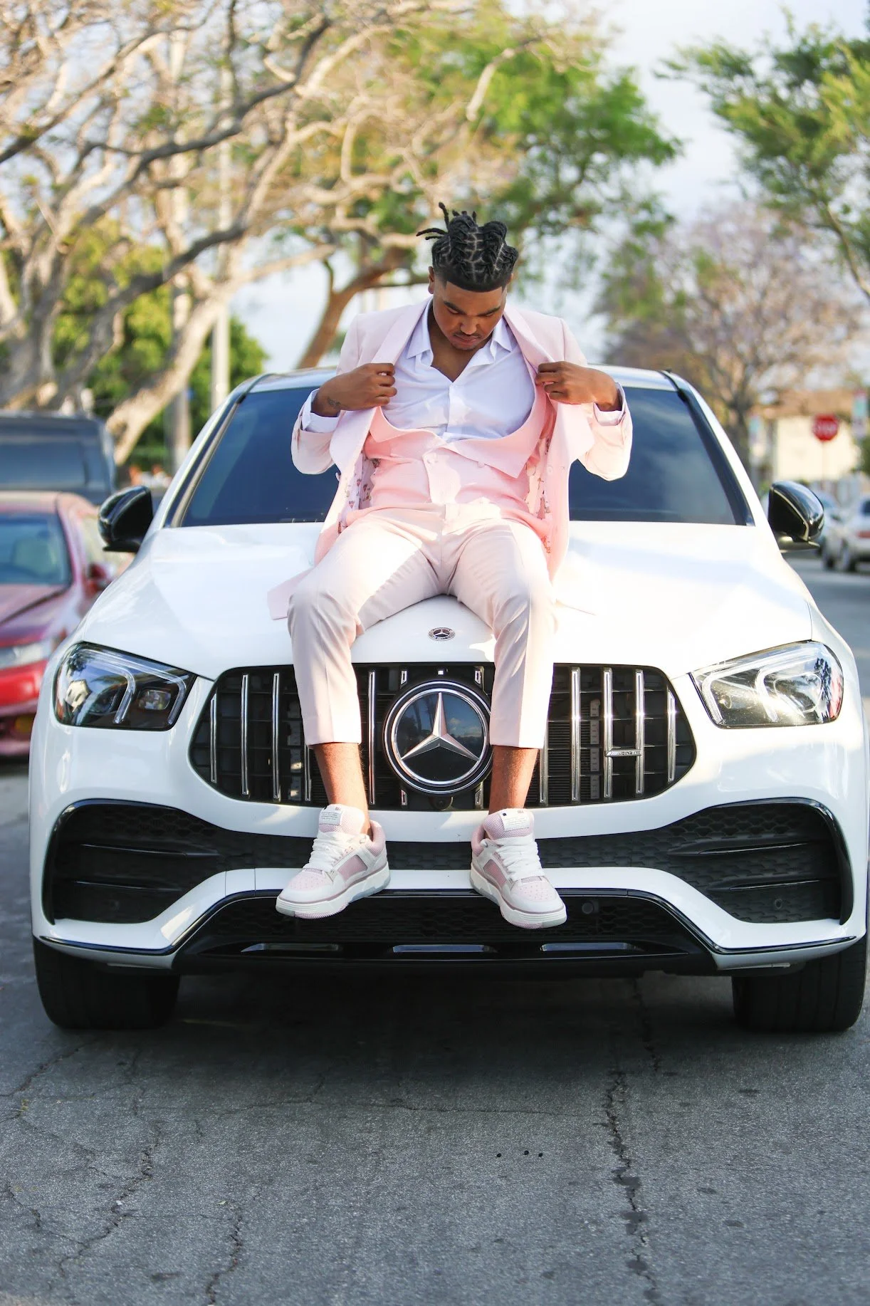 A man in a pink suit and sneakers sitting on the hood of a white Mercedes-Benz car on a city street.