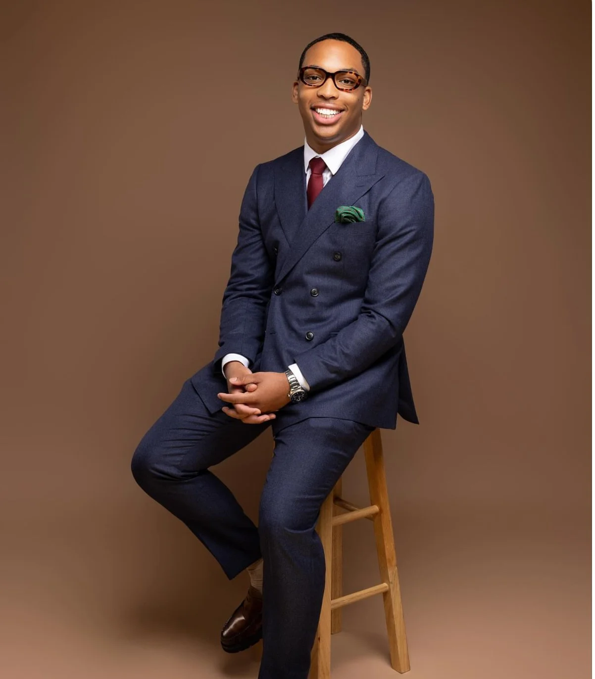 A man in a navy blue suit, white shirt, and red tie, sitting on a wooden stool against a brown background, smiling at the camera.