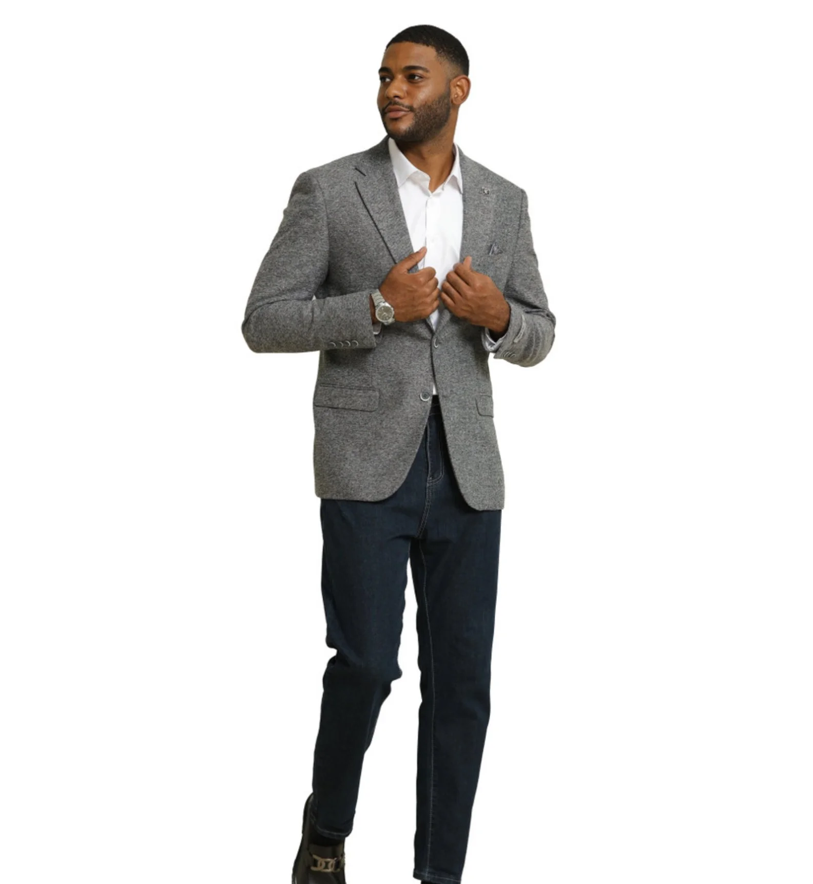 A man dressed in a gray blazer, white shirt, dark jeans, and black shoes, standing and adjusting his blazer.