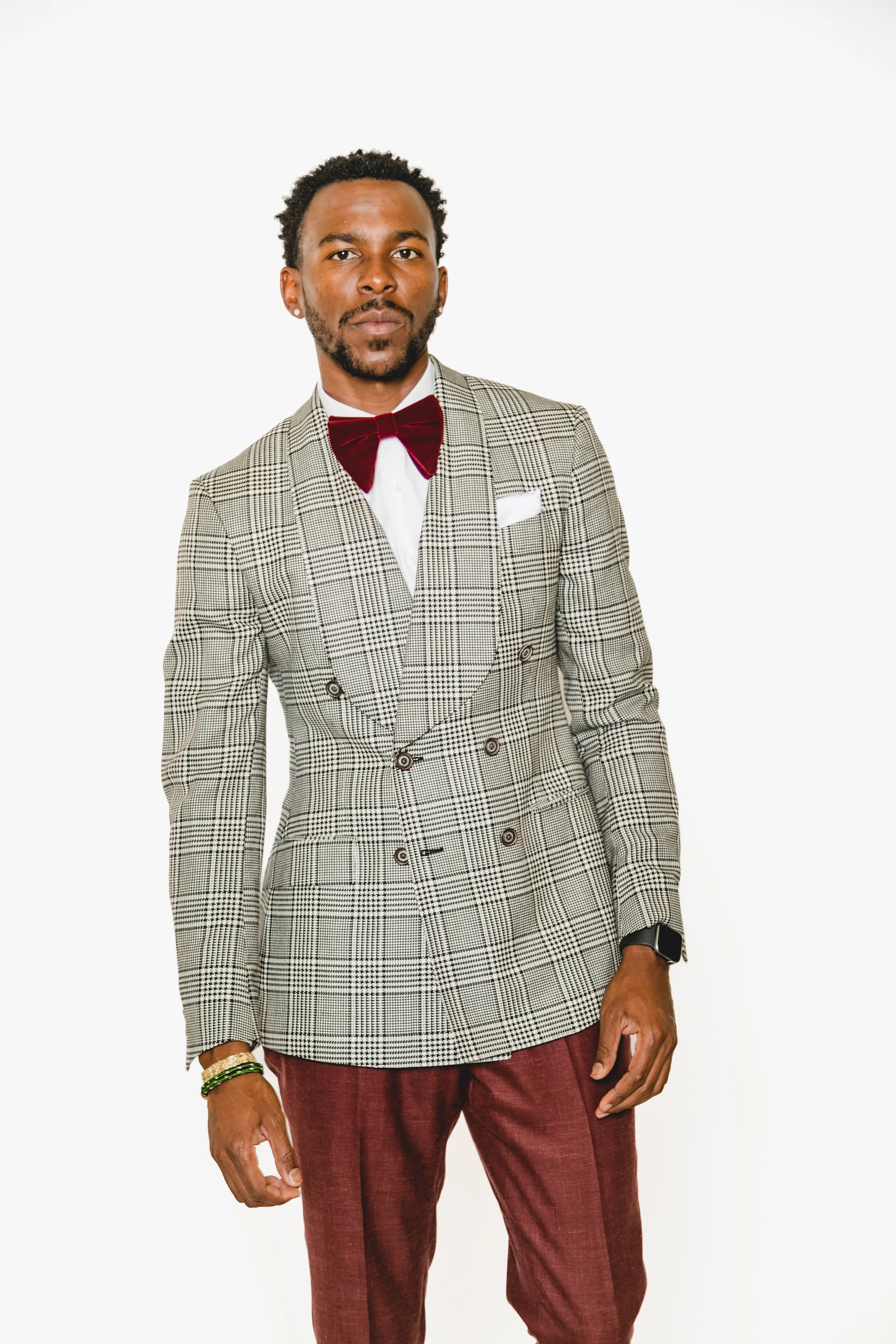 Man wearing a checked blazer with a red bowtie and maroon pants standing against a plain white background.