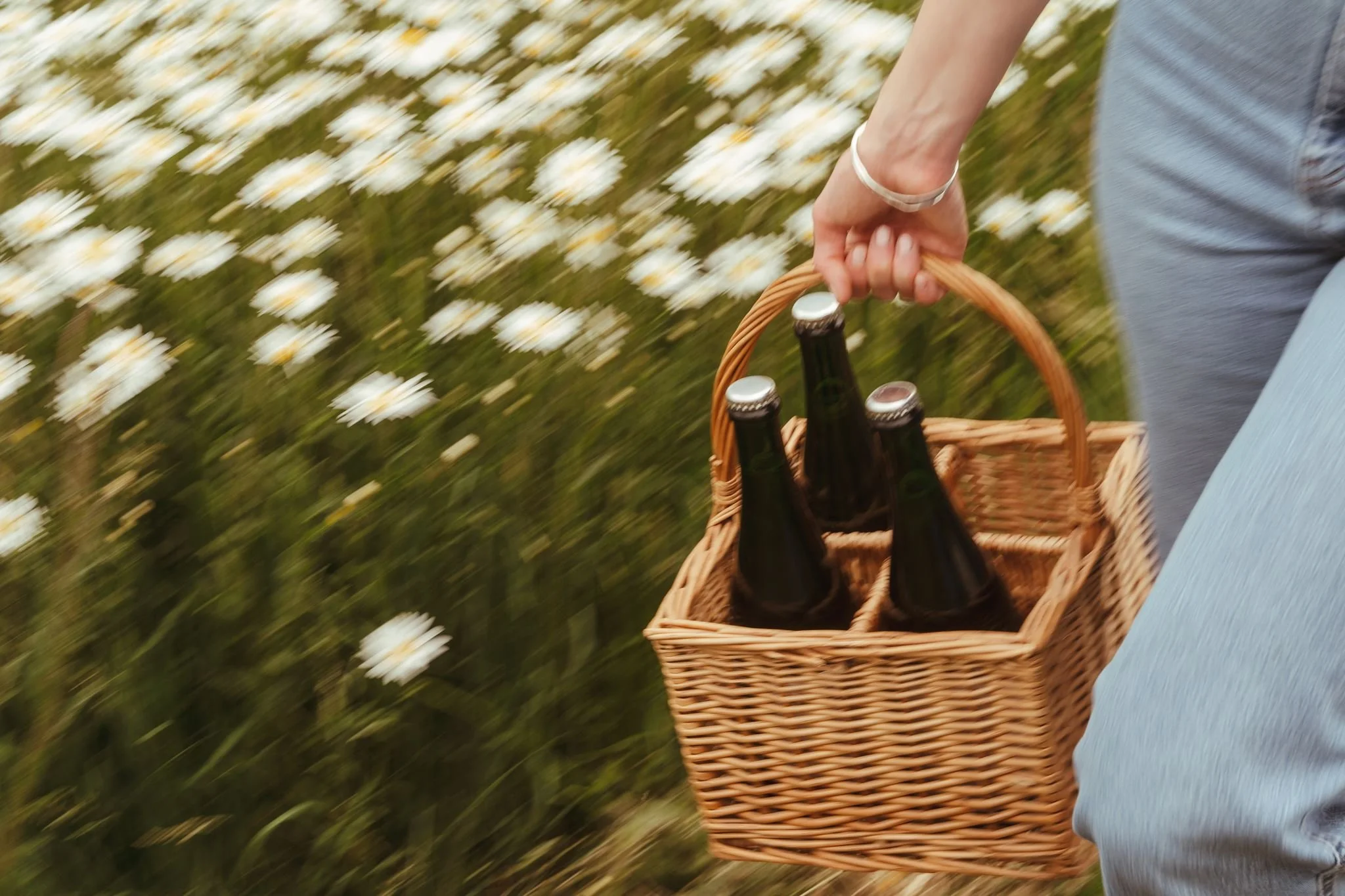 Person walking through a daisy field carrying a wicker basket with bottles.