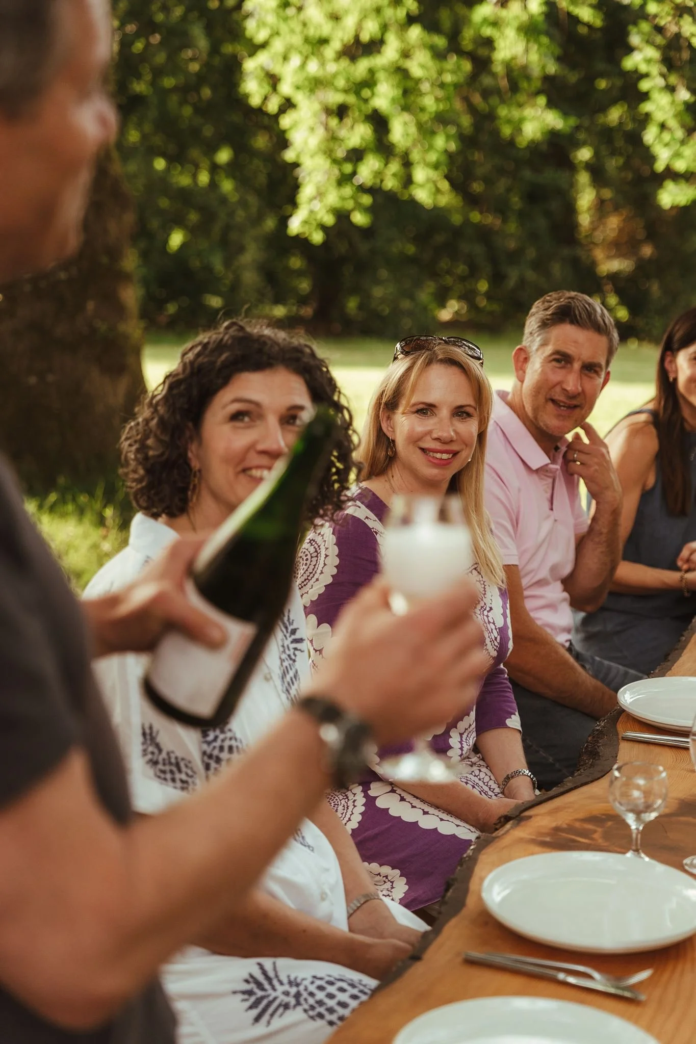 Group of people sitting at an outdoor dining table, smiling, with trees in the background, during daytime.