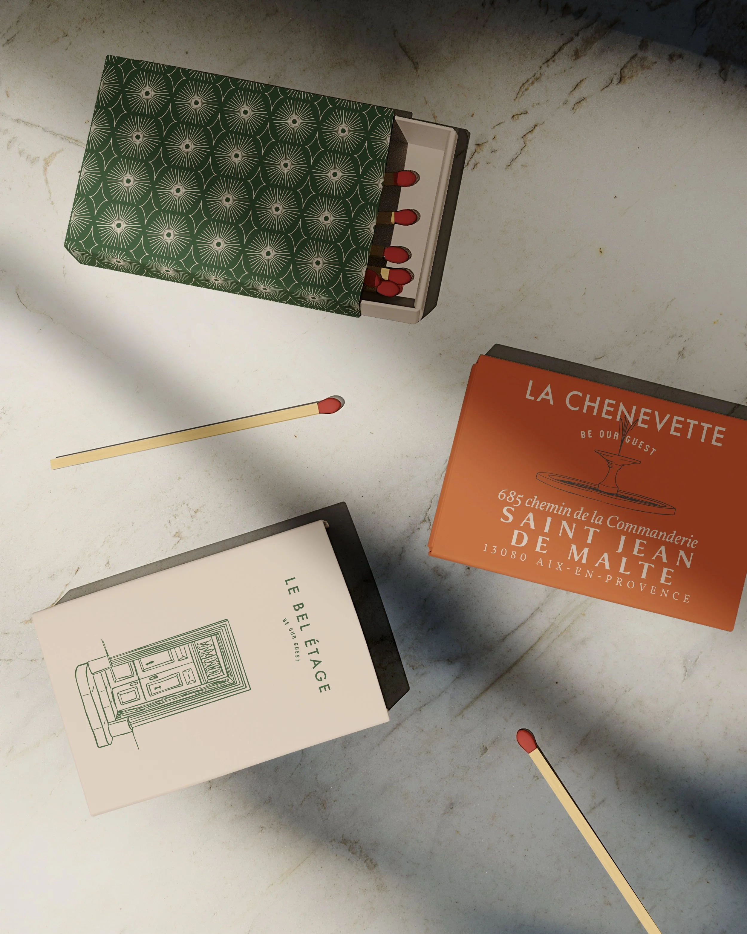 A decorative box with a geometric pattern containing matches with red tips, a white book with a green outline of furniture, and an orange menu card for a restaurant called La Chenette, seated on a marble countertop.
