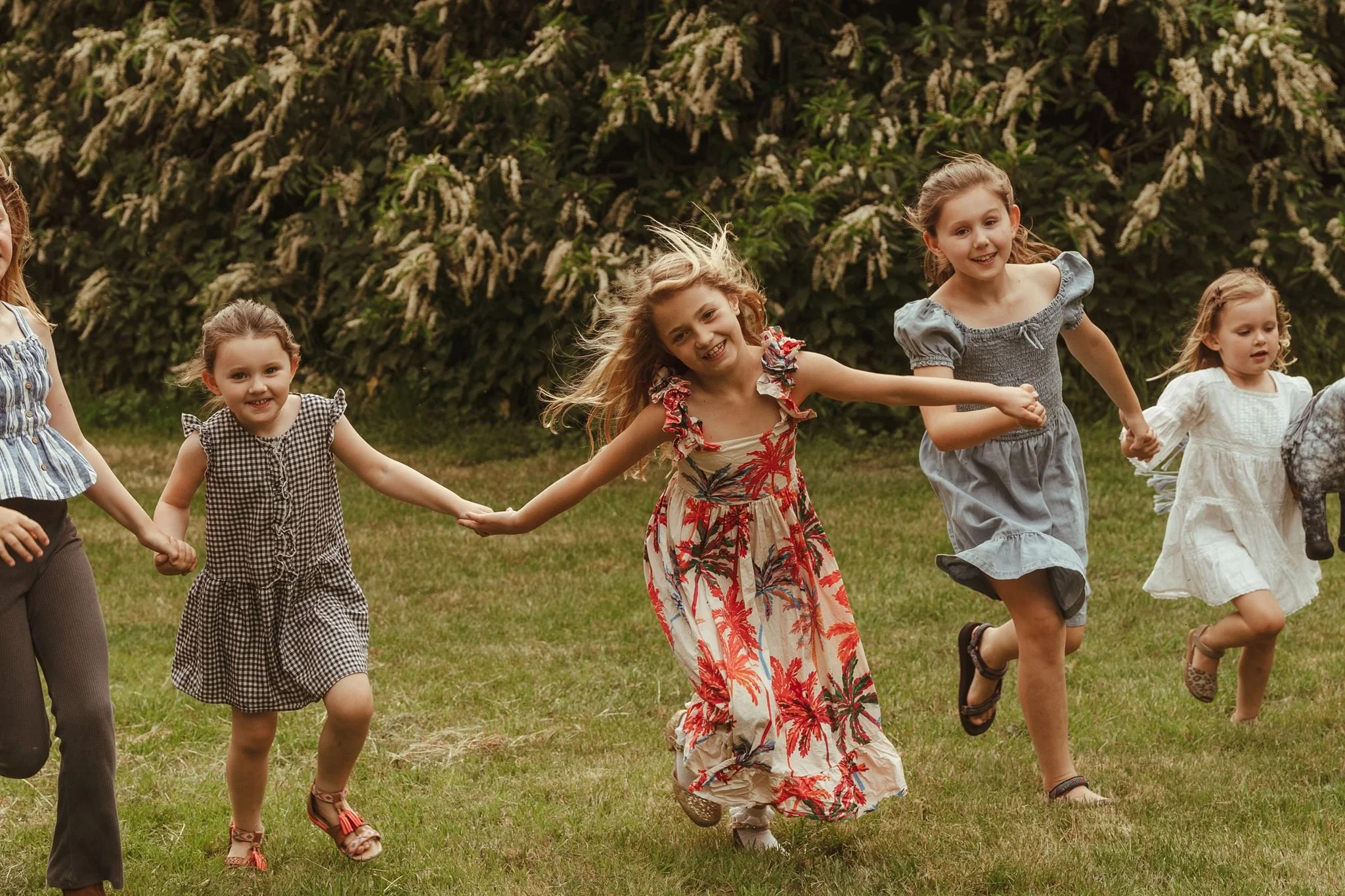 Group of young girls holding hands and running outdoors on grass during daytime, smiling and enjoying themselves.