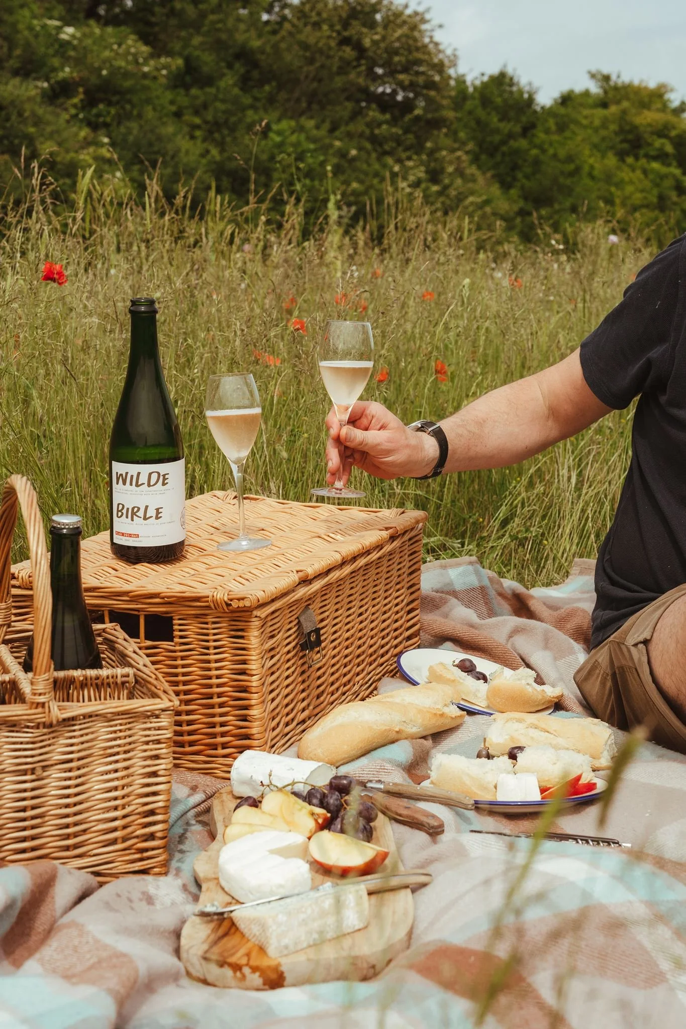 A person holding a glass of white wine outdoors during a picnic, with a wicker basket, a cheese platter, and bread on a blanket. There are wildflowers and grassy fields in the background.