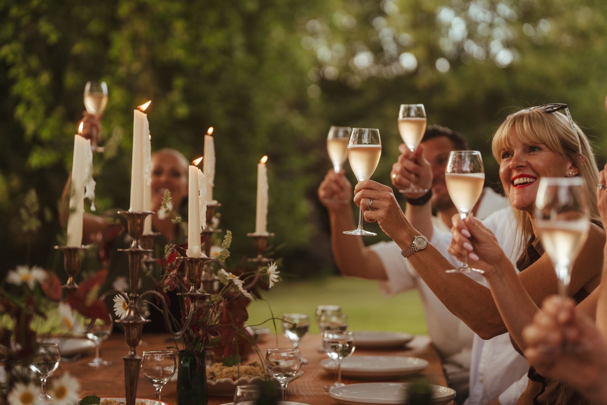 People raising glasses of champagne in a toast outdoors during sunset at a dinner party with candles and floral decorations.
