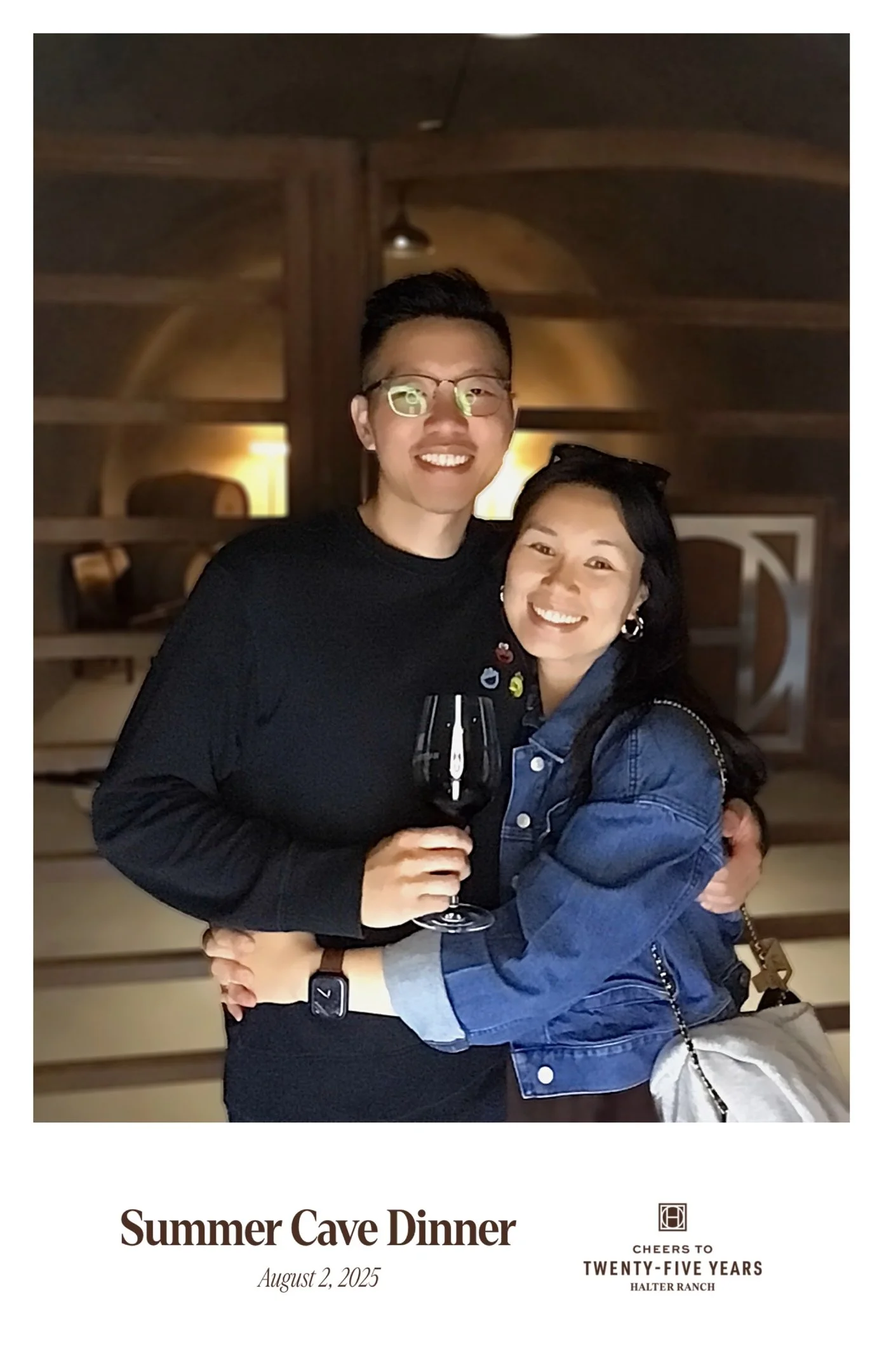 Two people hugging at a summer cave dinner, one holding a glass of red wine, smiling, inside a warmly lit cave or rustic setting.