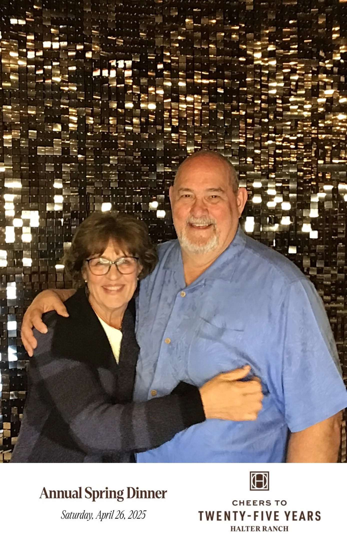 A smiling middle-aged man and woman hugging in front of a shiny, black sequin backdrop, with text indicating it's from the Annual Spring Dinner at Halter Ranch on April 26, 2025, celebrating 25 years.