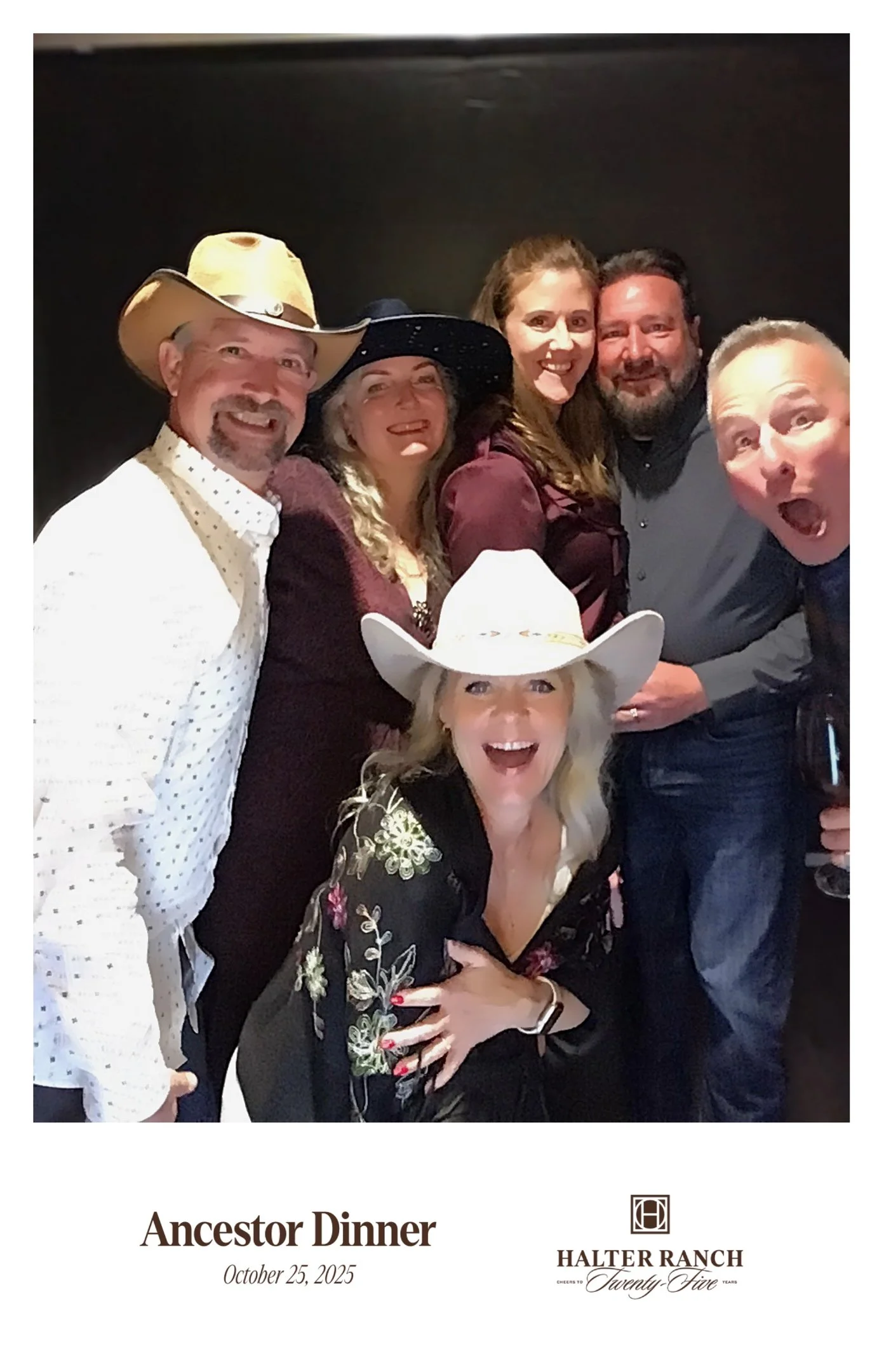 Group of people at an ancestor dinner event, some wearing cowboy hats, smiling and posing for a photo against a dark background.