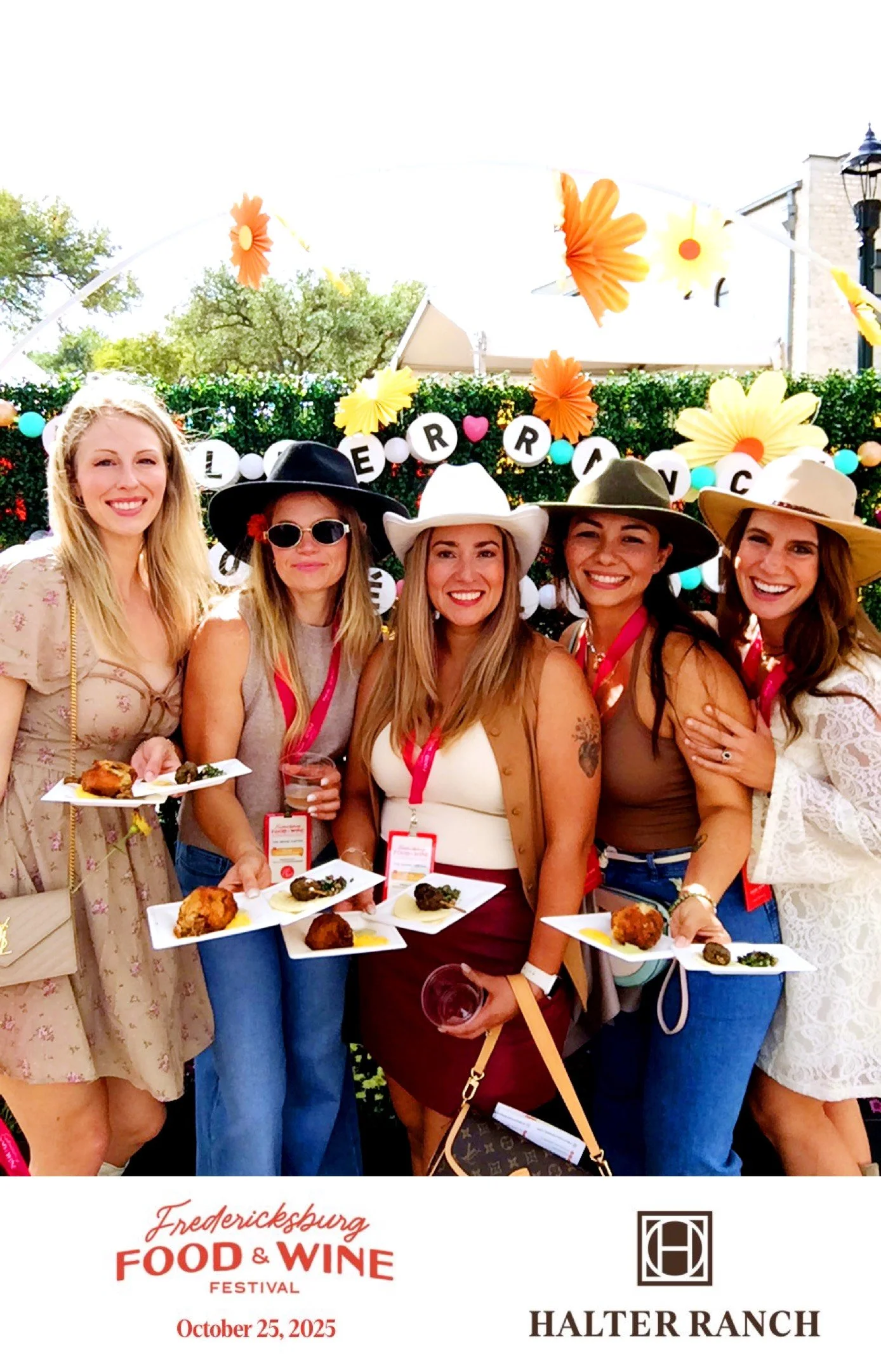 Five women wearing wide-brimmed hats at a food and wine festival, holding plates of appetizers, with a decorative flower backdrop and a sign reading 'Fredericksburg Food & Wine Festival October 25, 2025'.