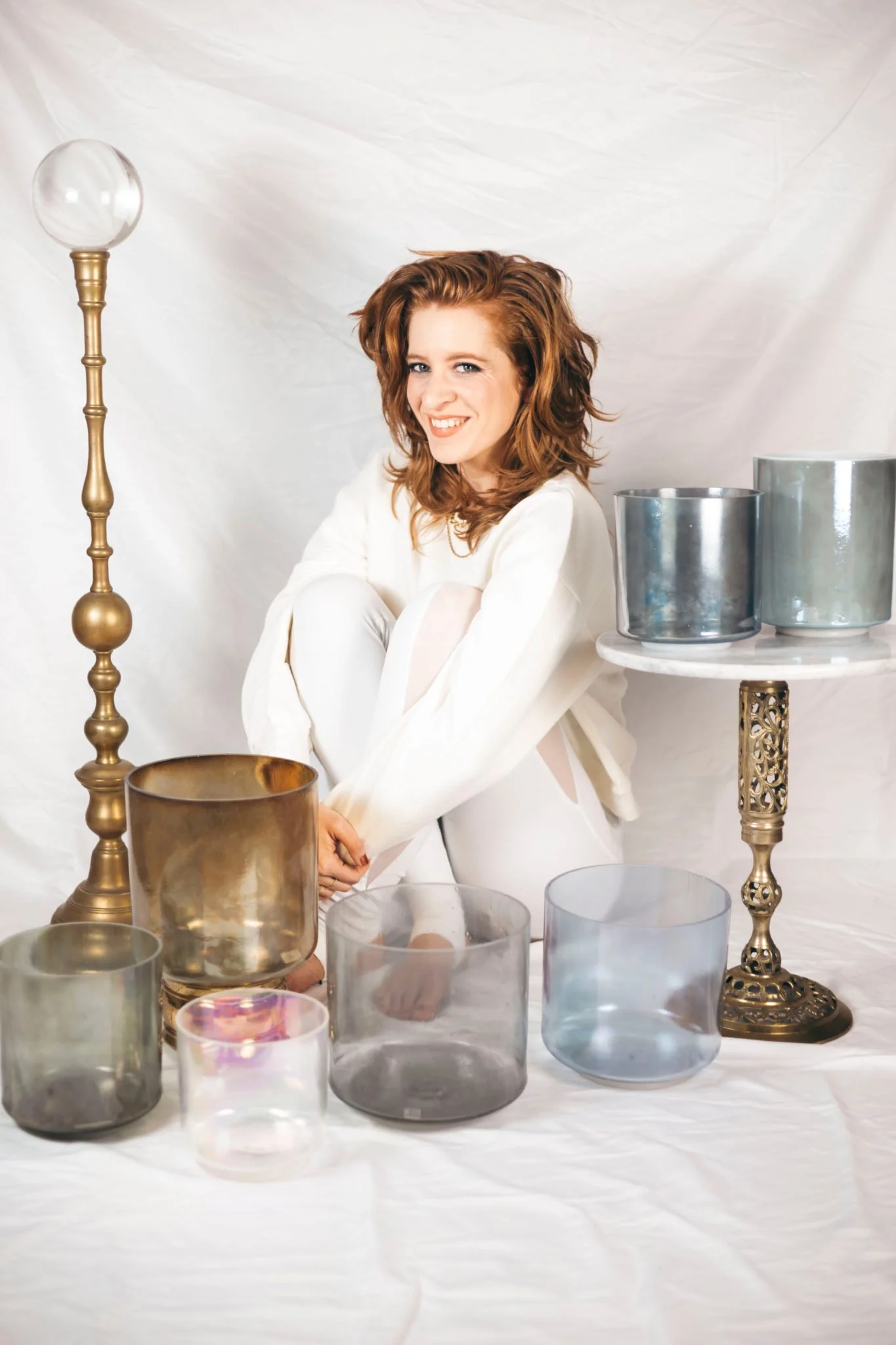 A smiling woman with red hair, wearing a white outfit, sitting on the floor surrounded by glass and metal decorative objects, including a tall brass lamp and various glass bowls, against a white backdrop.