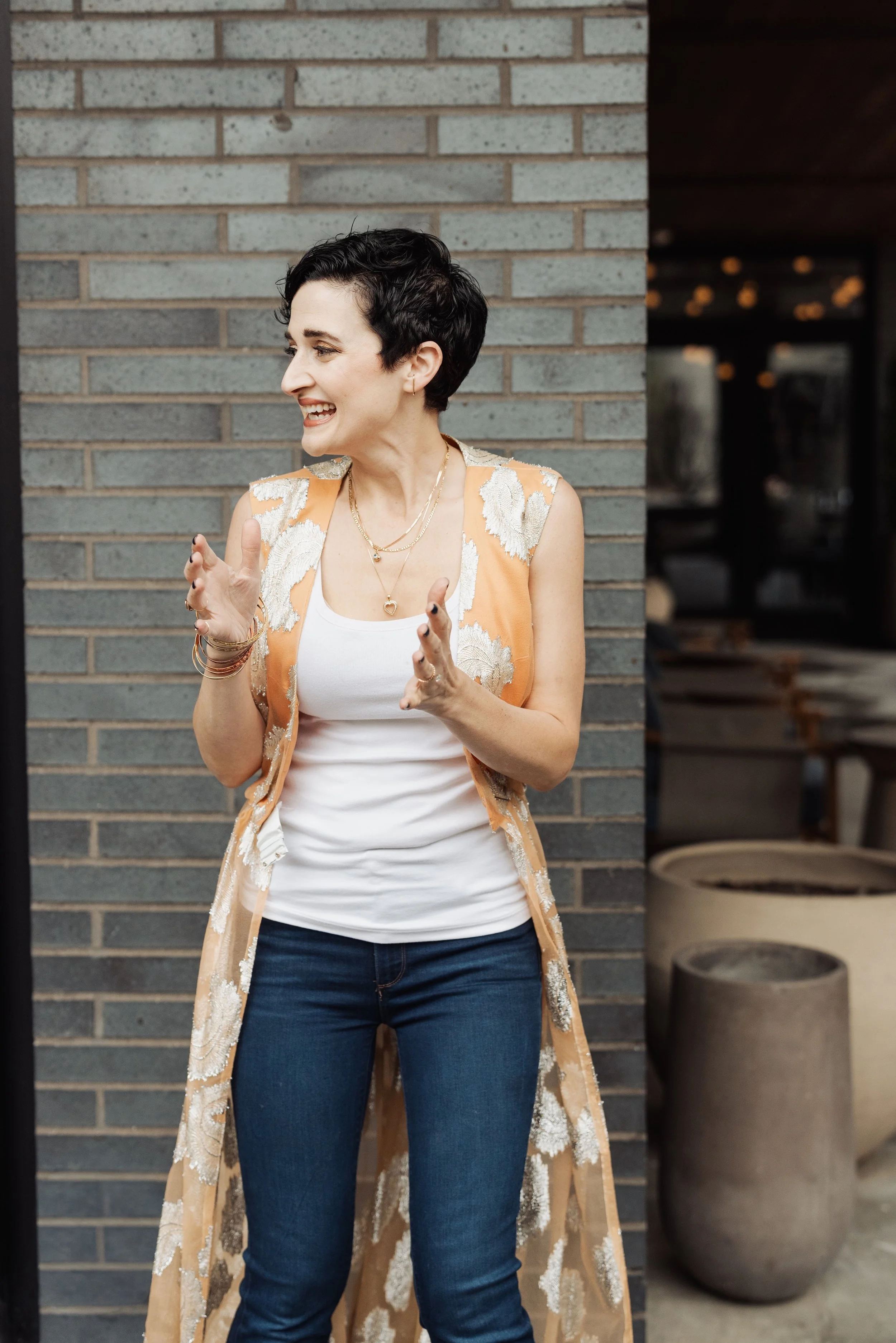 A woman with short dark hair smiling and talking, standing in front of a brick wall, wearing a white top, blue jeans, and a flowy beige and white patterned vest, accessorized with layered necklaces and bracelets.