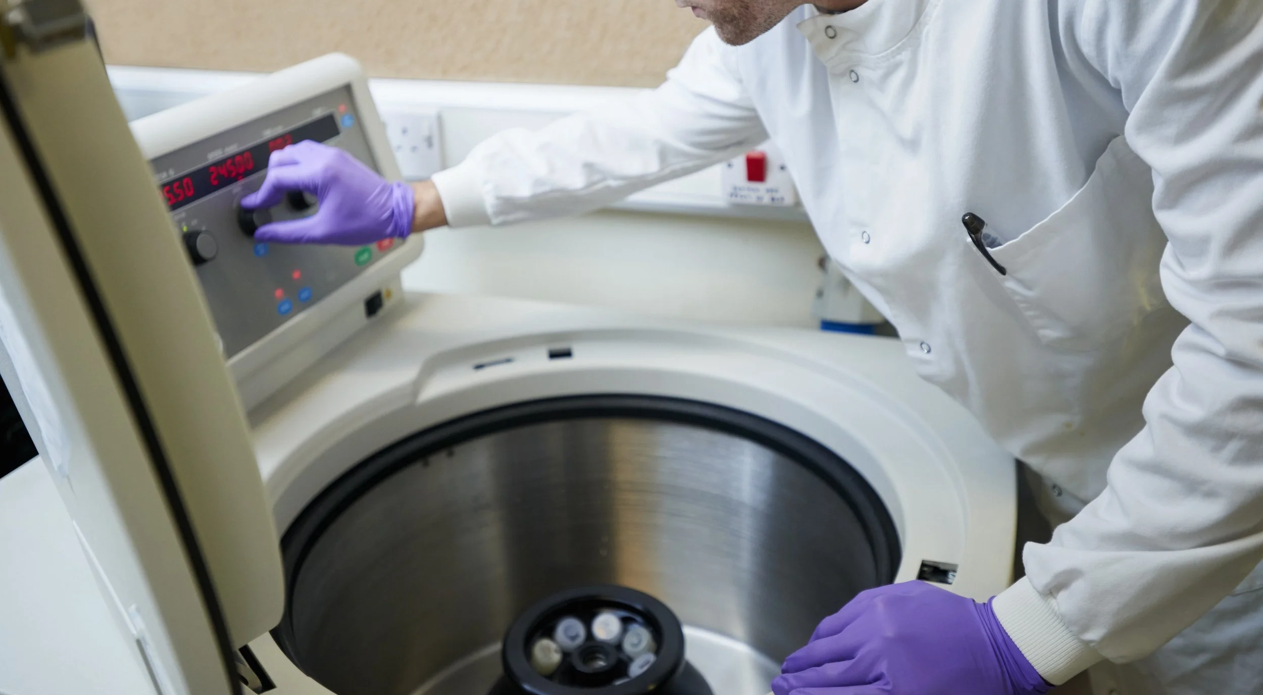 A scientist in a white lab coat and purple gloves operates a centrifuge in a laboratory setting.