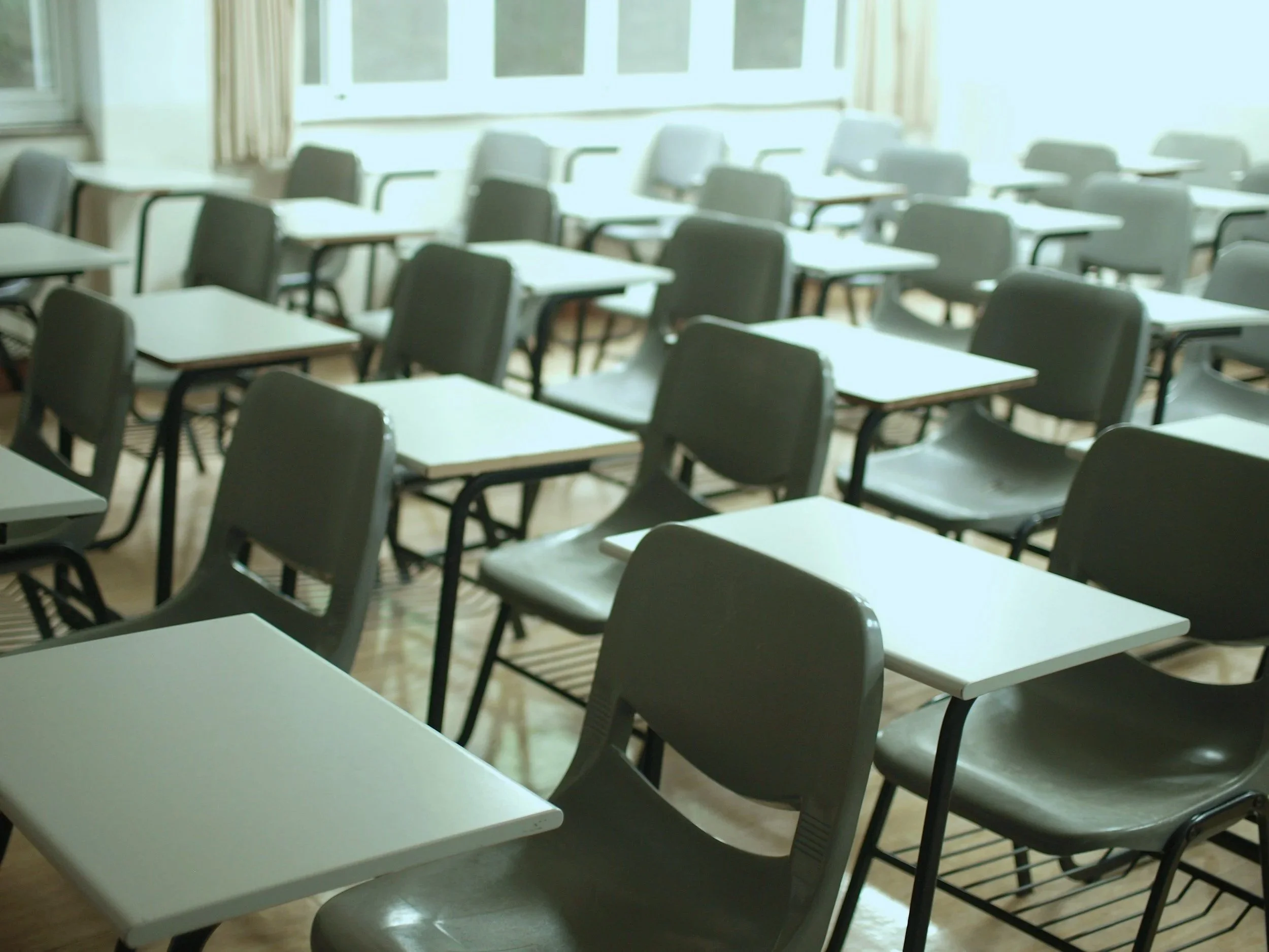 A classroom scene with rows of empty desks and green plastic chairs.