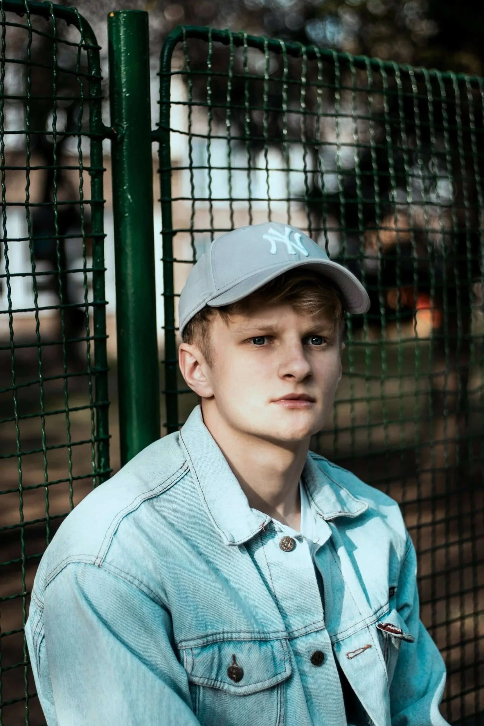 Young man wearing a light gray cap and denim jacket, leaning against a green metal fence.