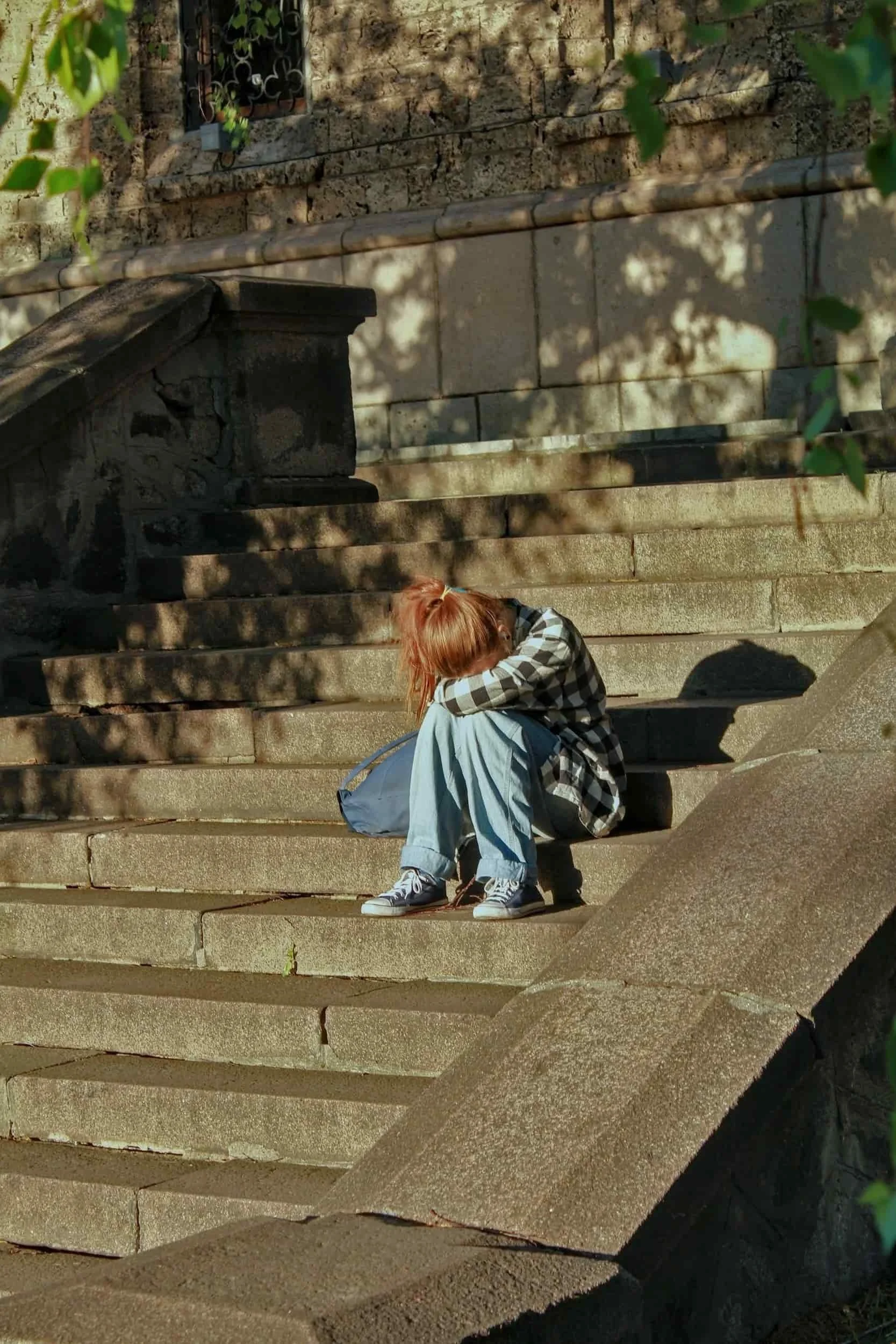 Person with red hair sitting on stone steps, covering their face with their arms, wearing a plaid shirt, jeans, and white sneakers, with green foliage and stone wall in the background.