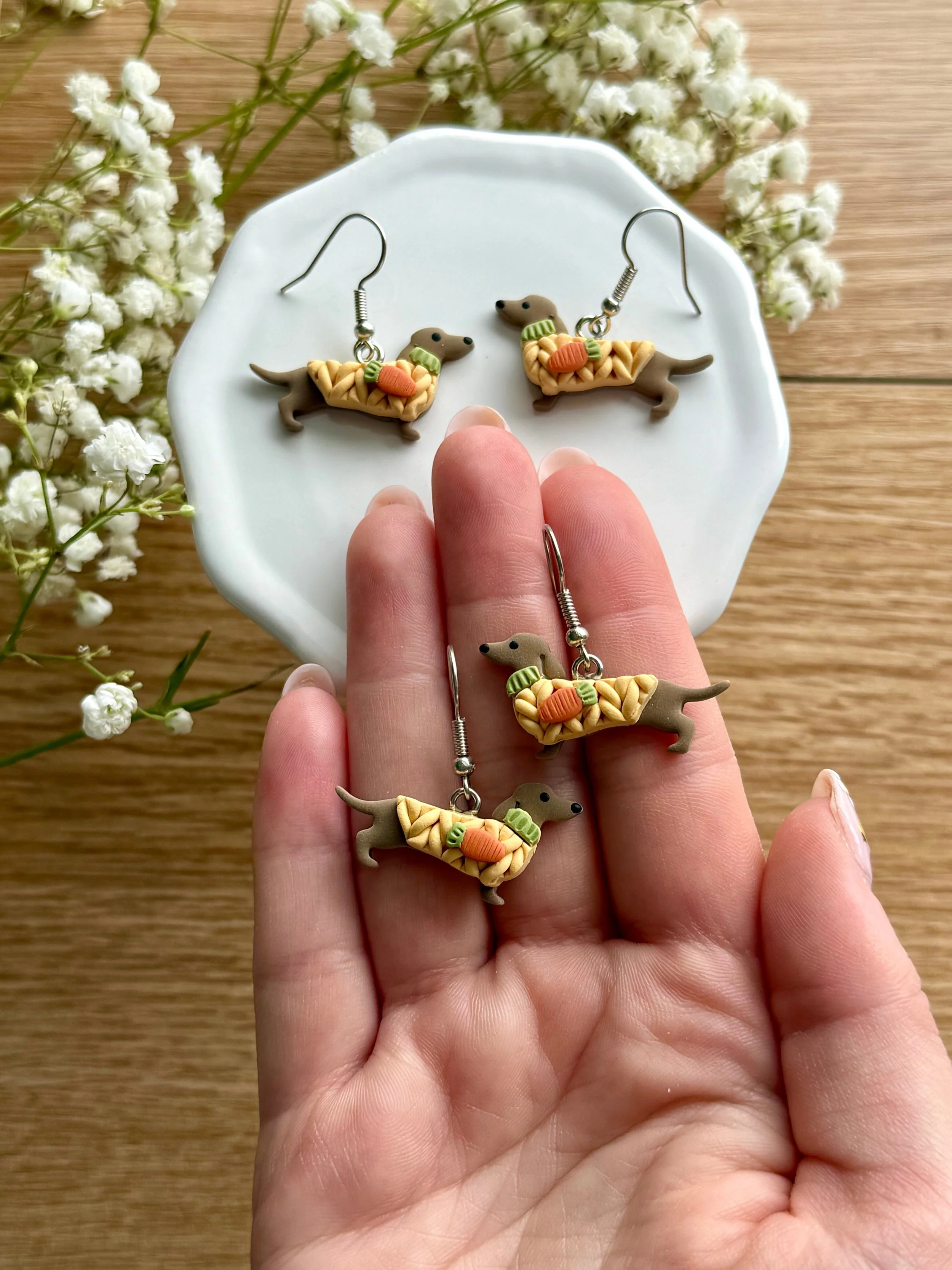 Leather dachshund-shaped jewelry set including earrings and a ring, decorated with small baguette and round beads in orange and green, displayed on a person's hand with a wooden background and white flowers.