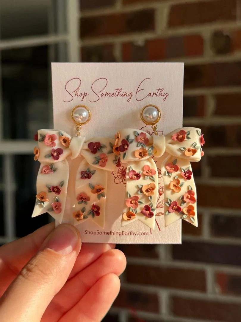 A pair of floral bow-shaped earrings with pearl studs attached to a card that says "Shop Something Earthy" and the website "ShopSomethingEarthy.com". The earrings are cream-colored with small pink, orange, and red flowers with green leaves.