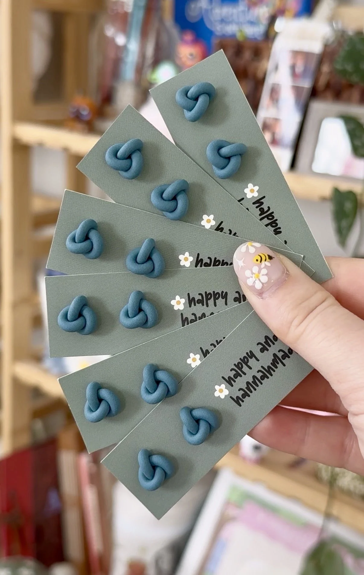 Hand holding five green cards with blue knot earrings and 'happy and' printed on them. The person's finger has white nail polish with a bee and flower design. The background appears to be a store with shelves and pictures.