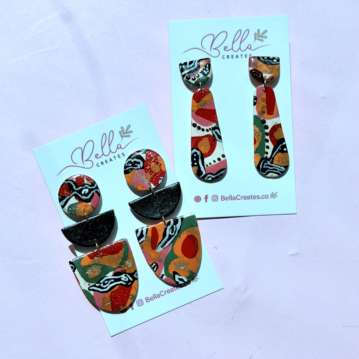 Two pairs of colorful, abstract-patterned earrings displayed on Bella Creates cards, set against a light pink background.