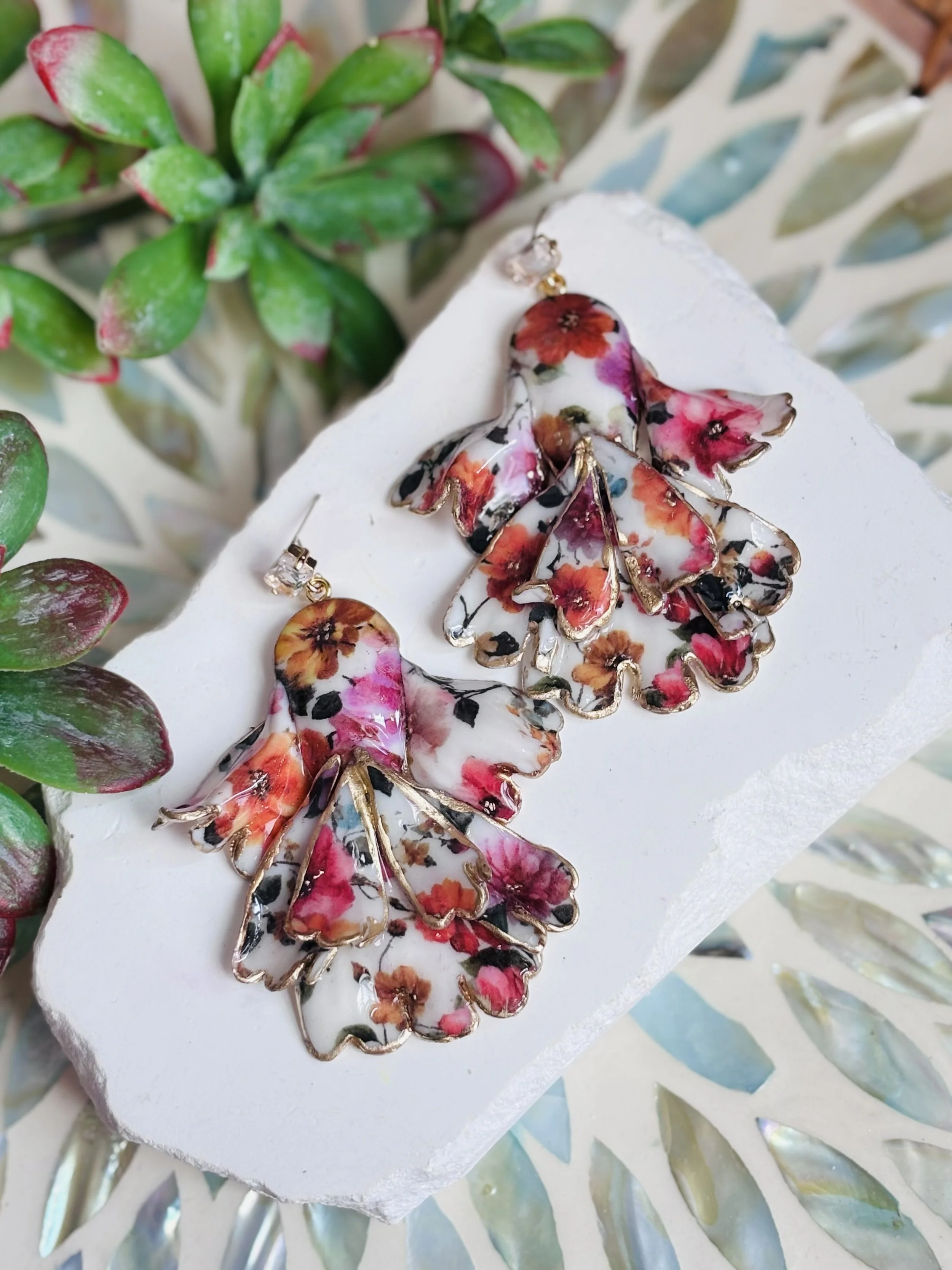By Hollee from GlamPop Designs, Colorful floral earrings with gold accents displayed on a white surface, surrounded by green succulent plants.