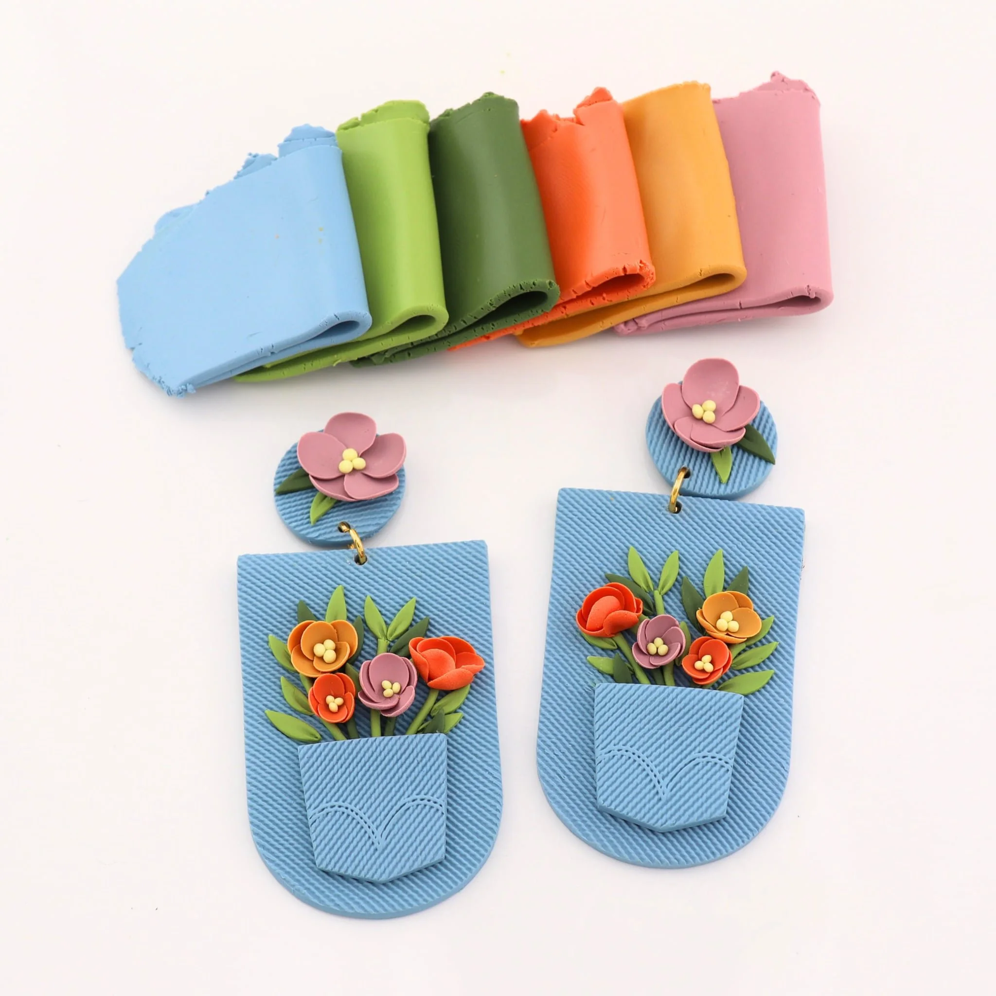 Pair of colorful earrings with pink flower top and blue base with orange and pink flowers and green leaves, in front of a row of rolled up colorful clay or fabric in blue, green, dark green, orange, yellow, and pink.
