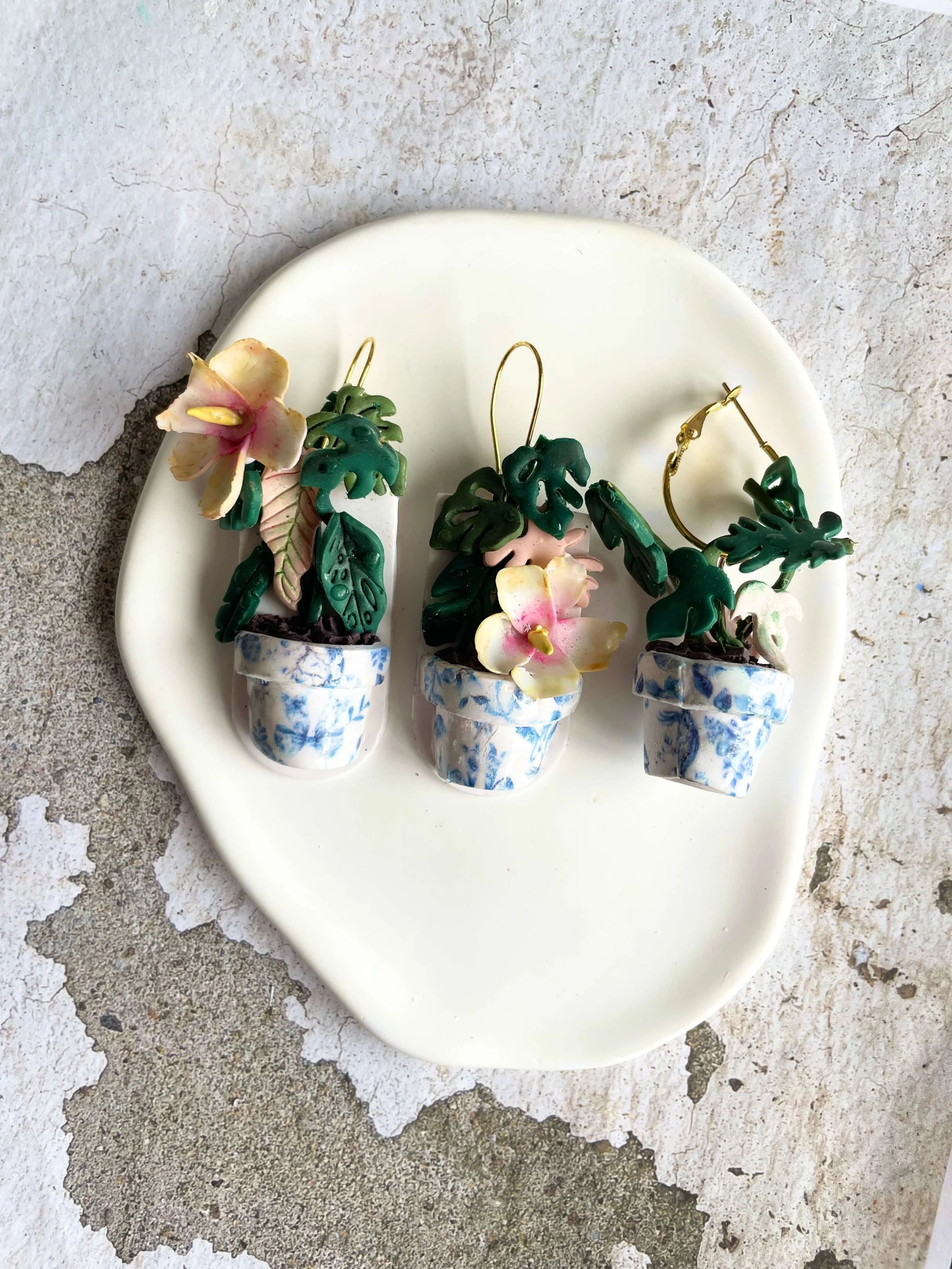 Three flower-shaped earrings with green leaf designs in blue and white pots, placed on an oval white ceramic dish on a textured, weathered surface.