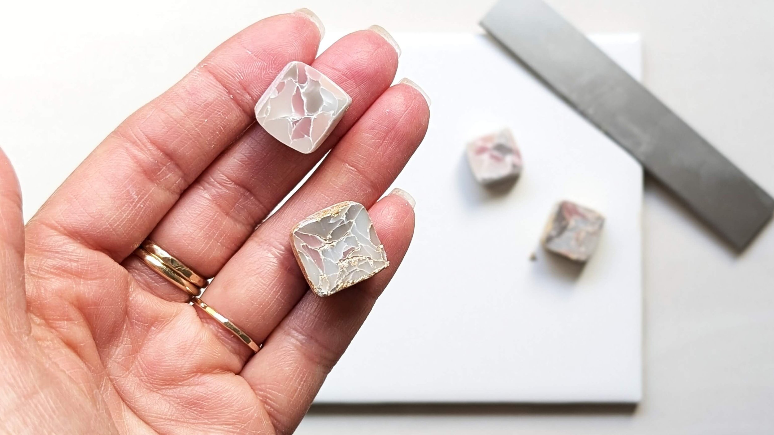 Hand holding two square marble tiles with white and pink veining, with a few similar tiles on a white surface nearby.