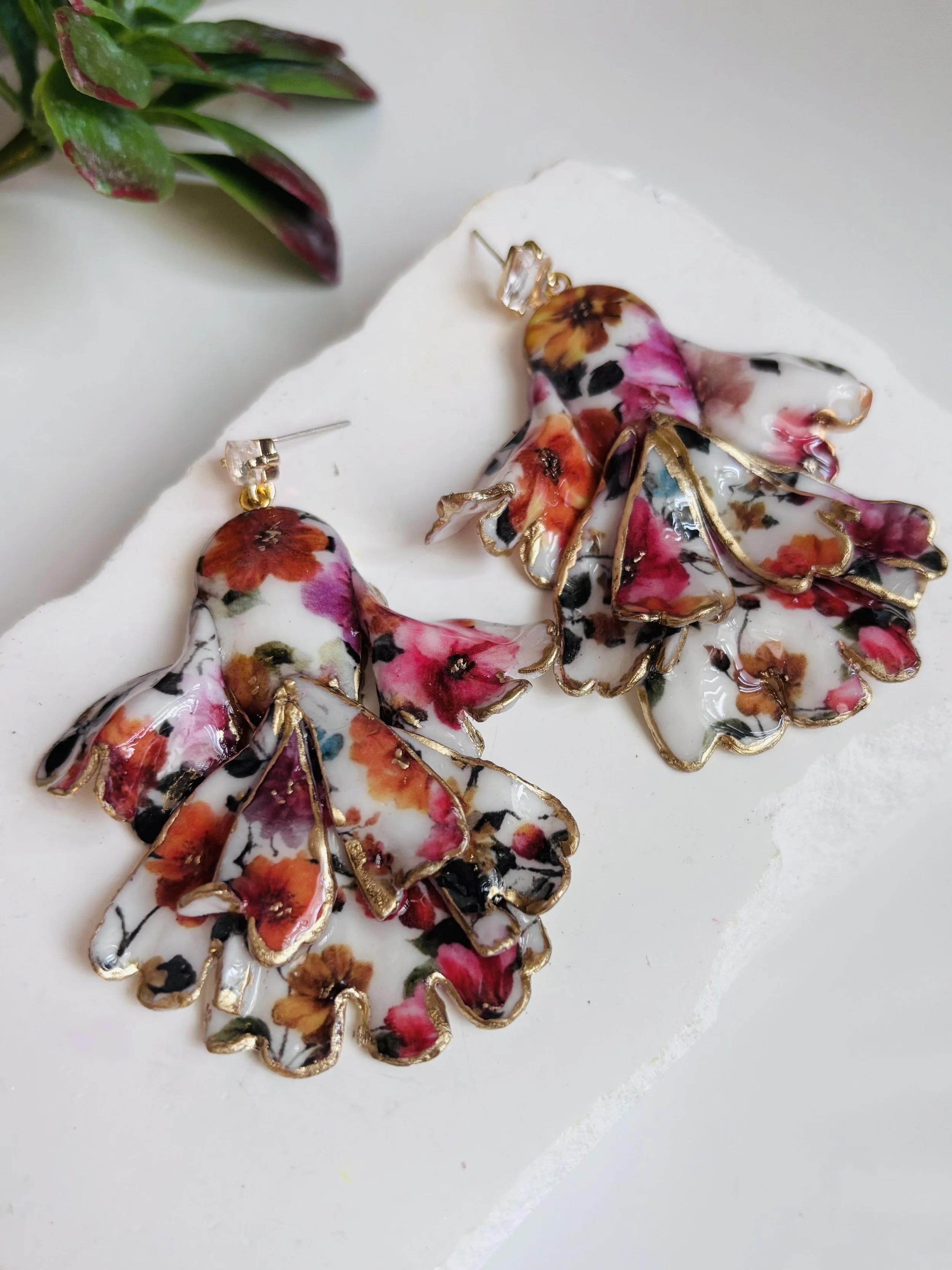 Floral butterfly-shaped earrings with gold trim on a white surface with a green plant in the background.
