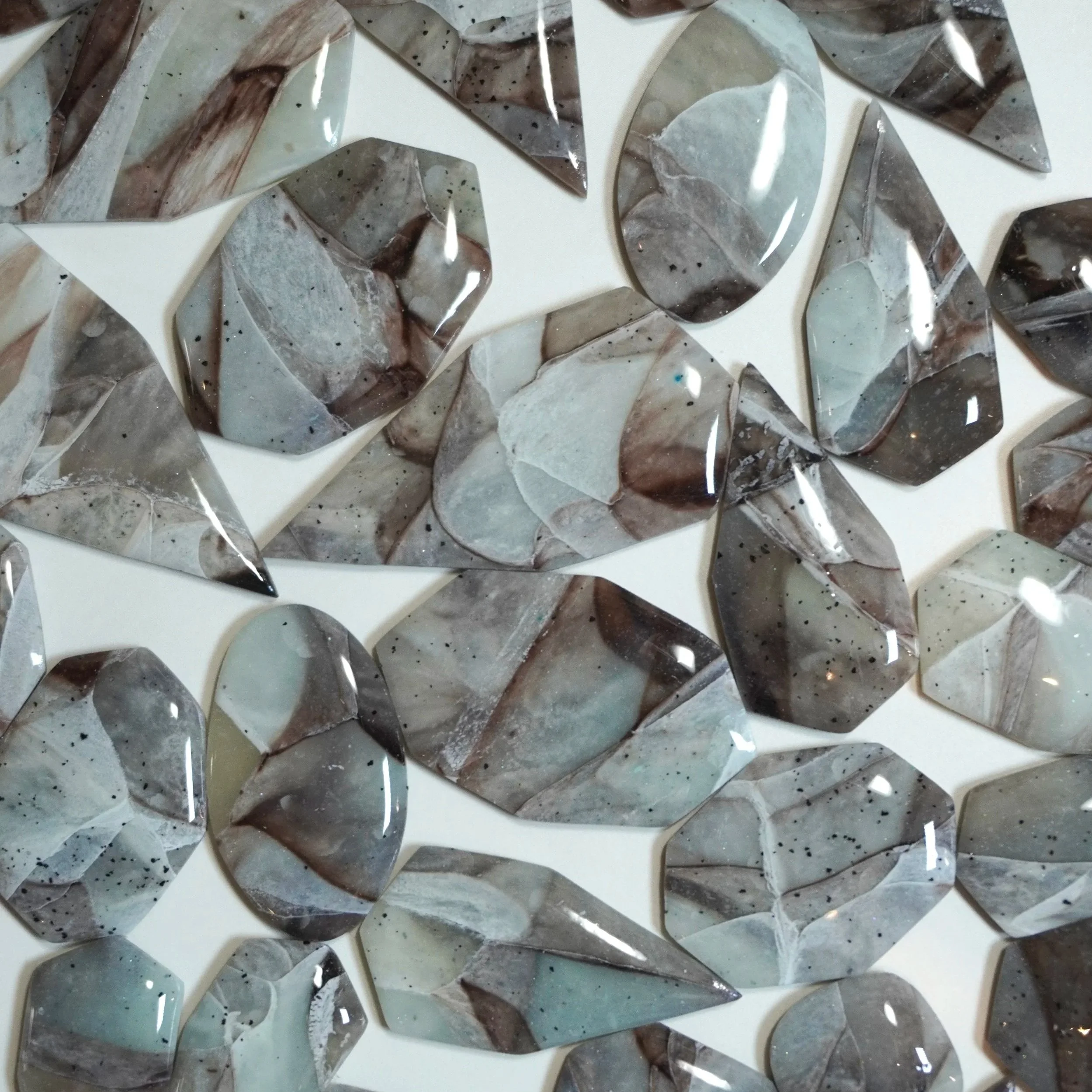 Collection of polished, irregularly shaped gemstones with gray, white, and brown swirling patterns and small black specks on a white surface.
