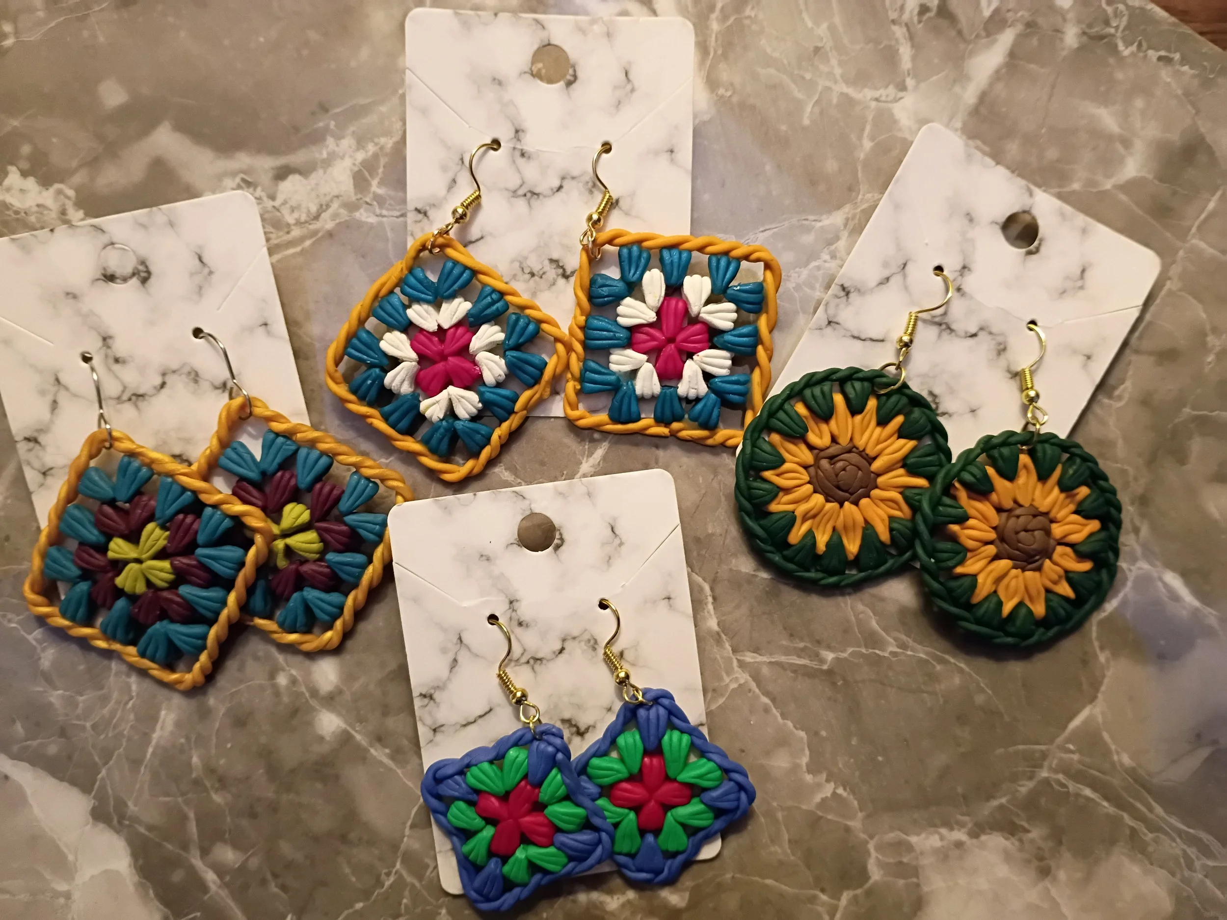 Colorful handcrafted earrings with floral and geometric patterns displayed on white marble-patterned cards, set on a marble surface.