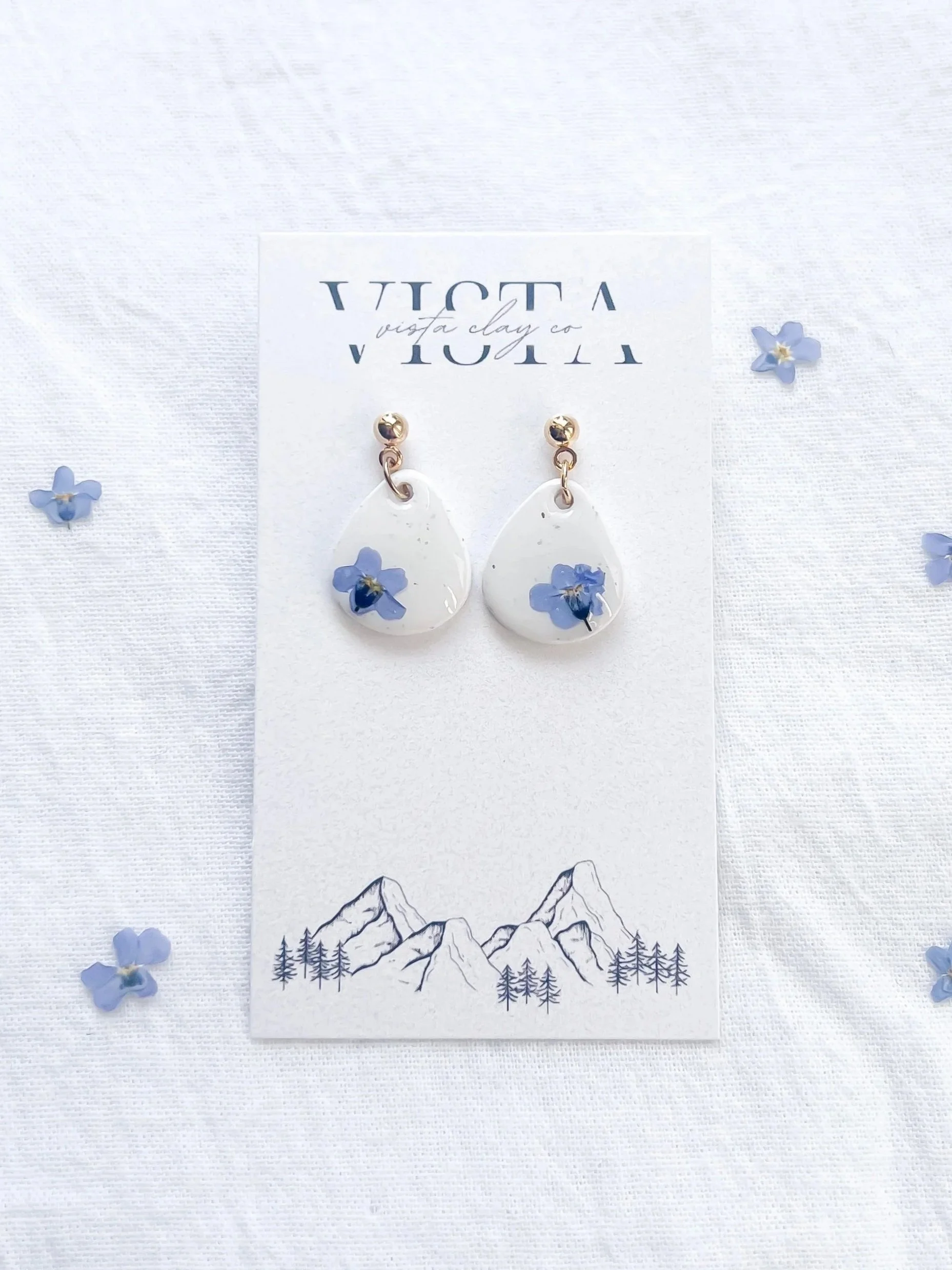 White earrings with blue flowers, displayed on a white card with mountain drawing, surrounded by small blue flower confetti.