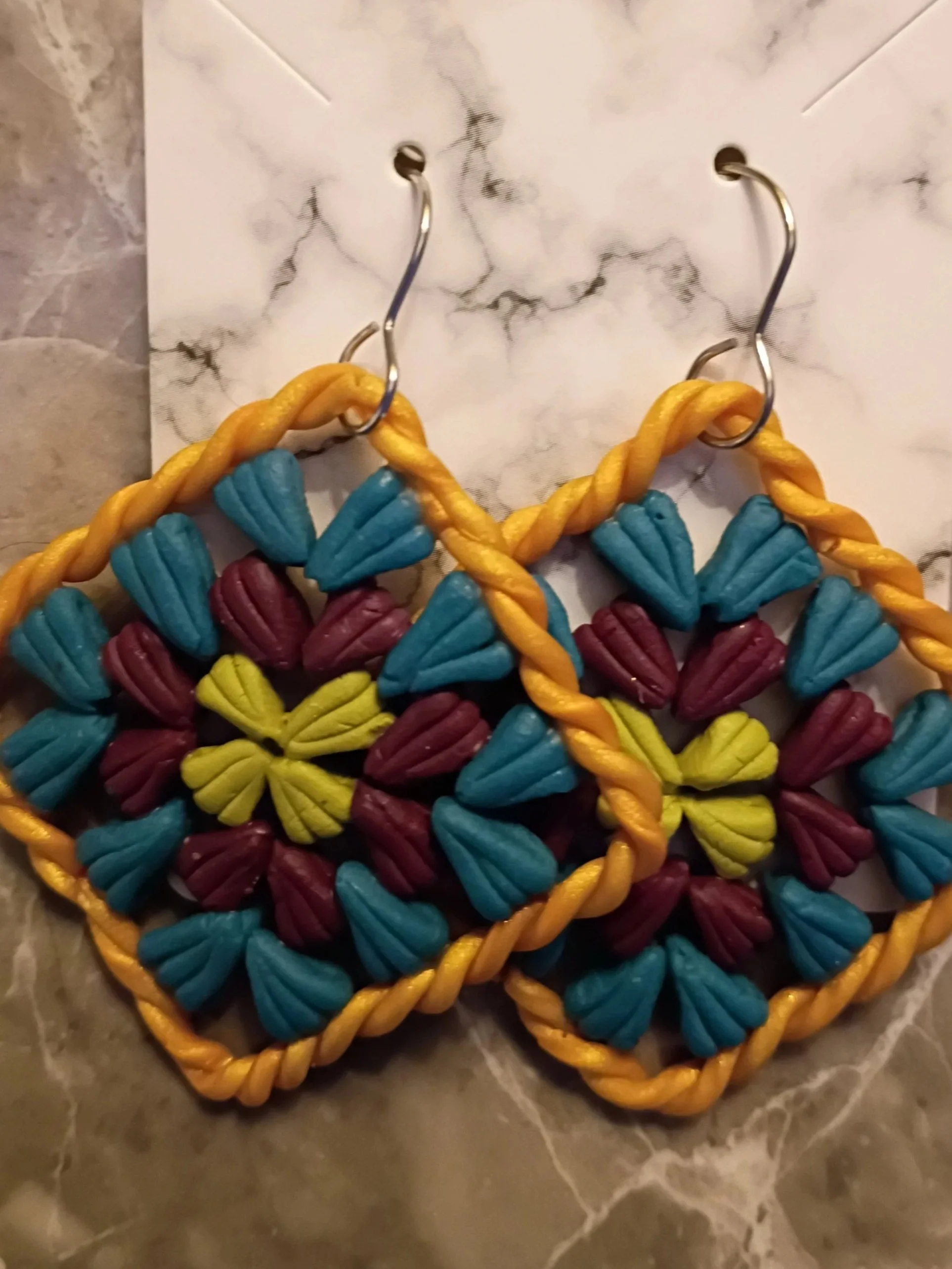 Colorful woven earrings with a heart shape, featuring blue, maroon, yellow, and teal sections, hanging from hooks on a marble surface.