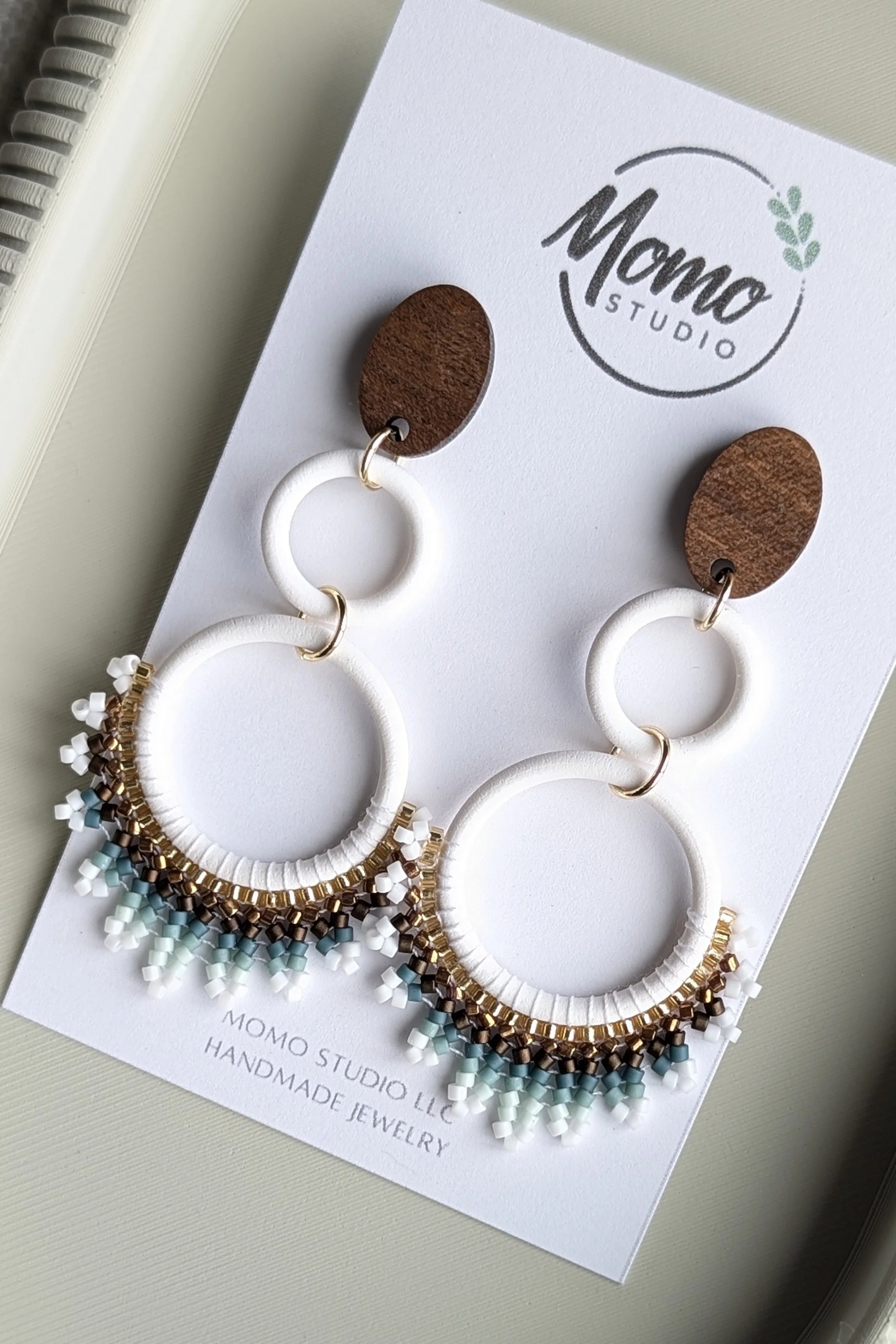 Beaded earrings with white, gold, brown, and blue beads hanging from circular white and brown elements, displayed on MOMO Studio handmade jewelry card.