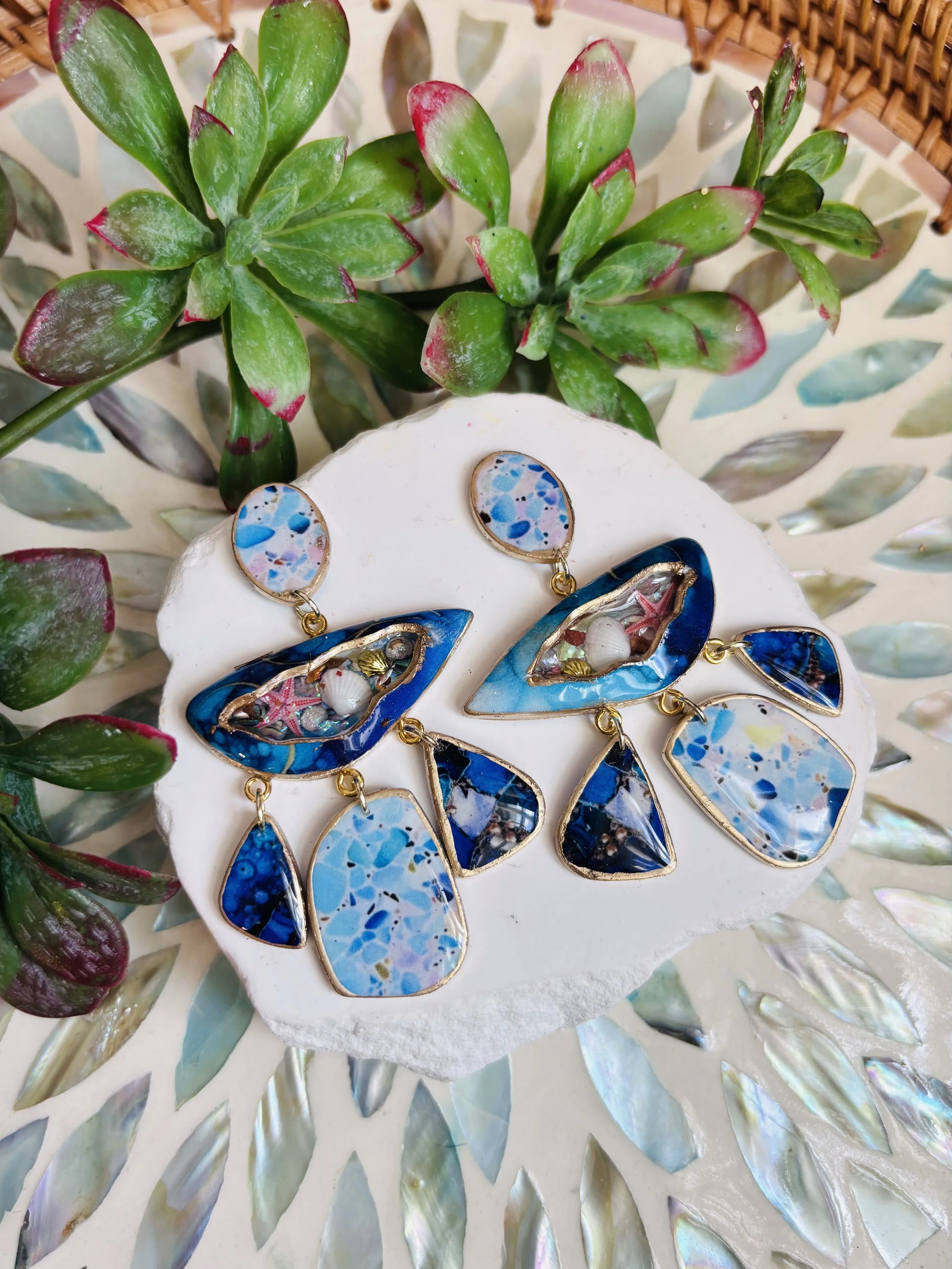 Colorful earrings with blue and pink abstract patterns and small decorative elements, displayed on a white surface with green plants in the background.