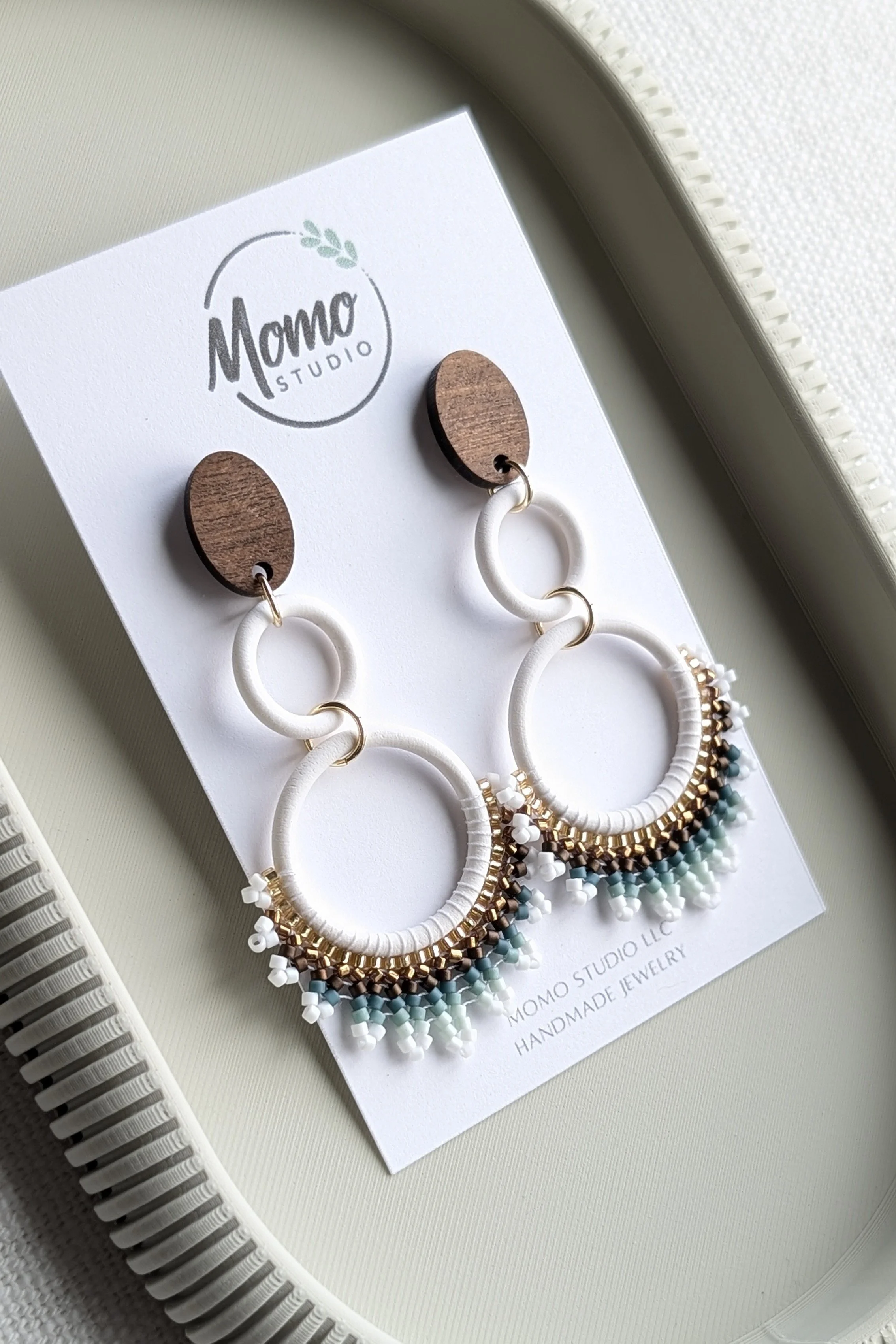 Pair of handmade hoop earrings with wood, white, gold, and teal beads, displayed on a white card from Momo Studio with a printed logo, on a white tray.
