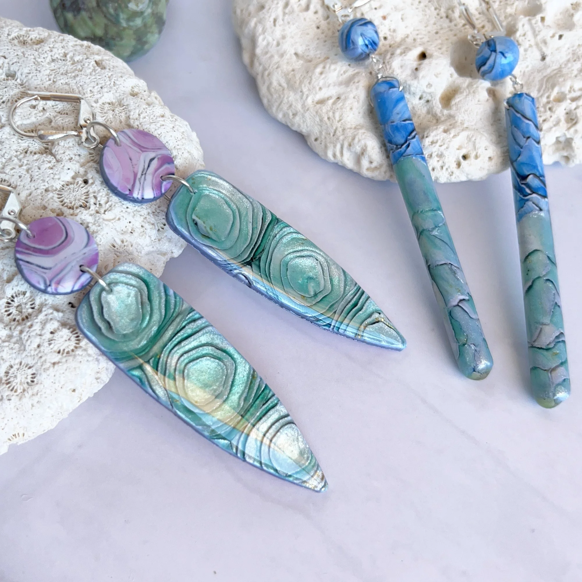 Colorful handmade jewelry, including earrings and bracelets, displayed on white lava rocks. The jewelry features abstract swirl patterns in pastel shades of purple, green, and blue with metallic accents.